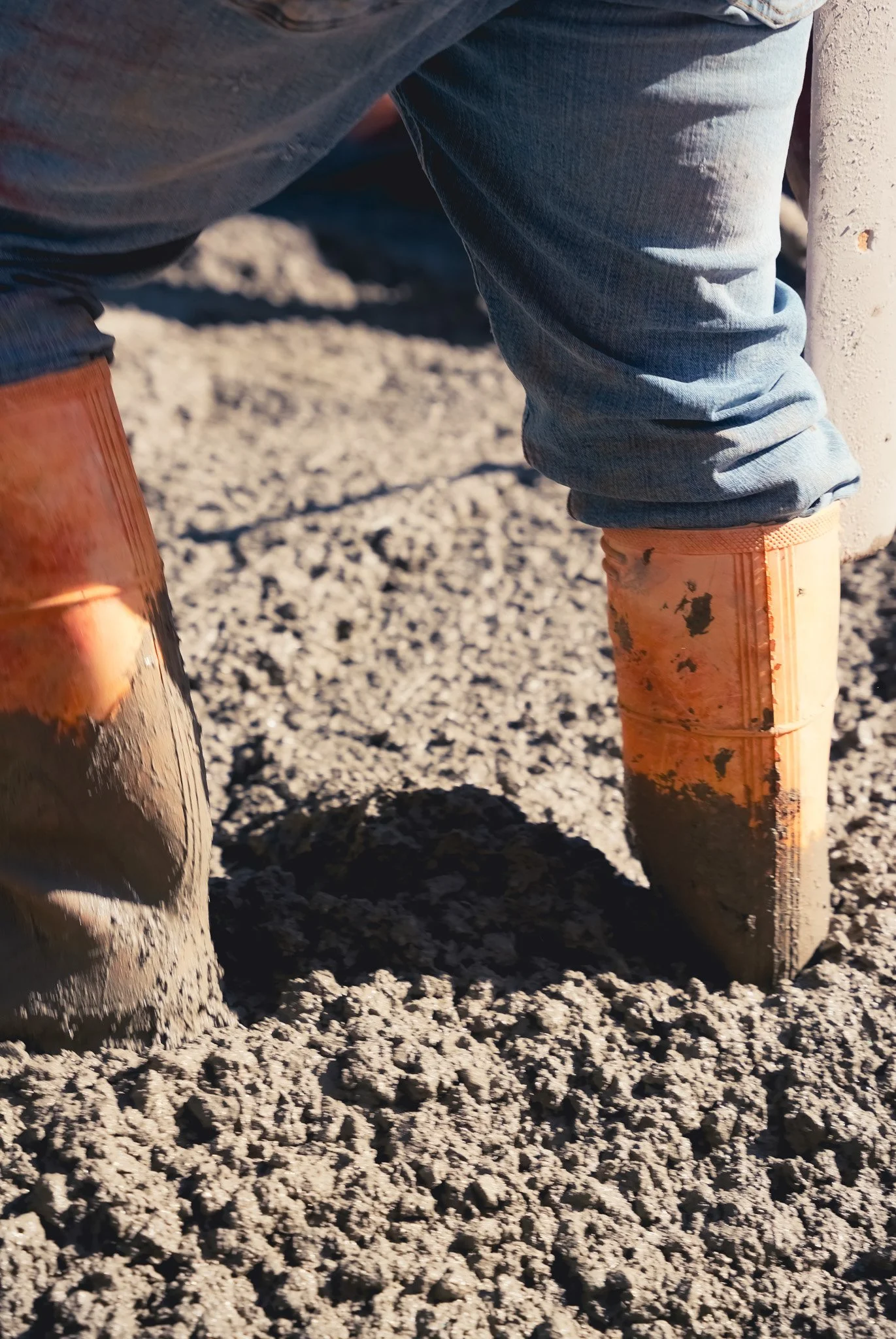 Close-up of a person wearing blue jeans and orange rubber boots standing on wet gravel or dirt ground.
