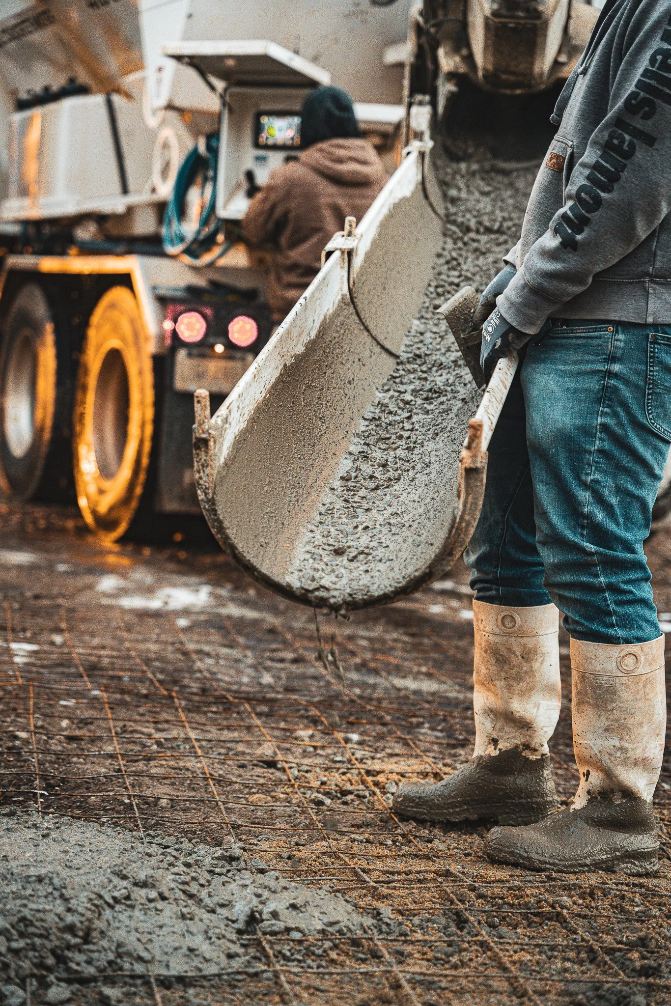 Worker spreading wet concrete on a construction site using a large shovel, with another worker in the background operating a concrete mixer truck.