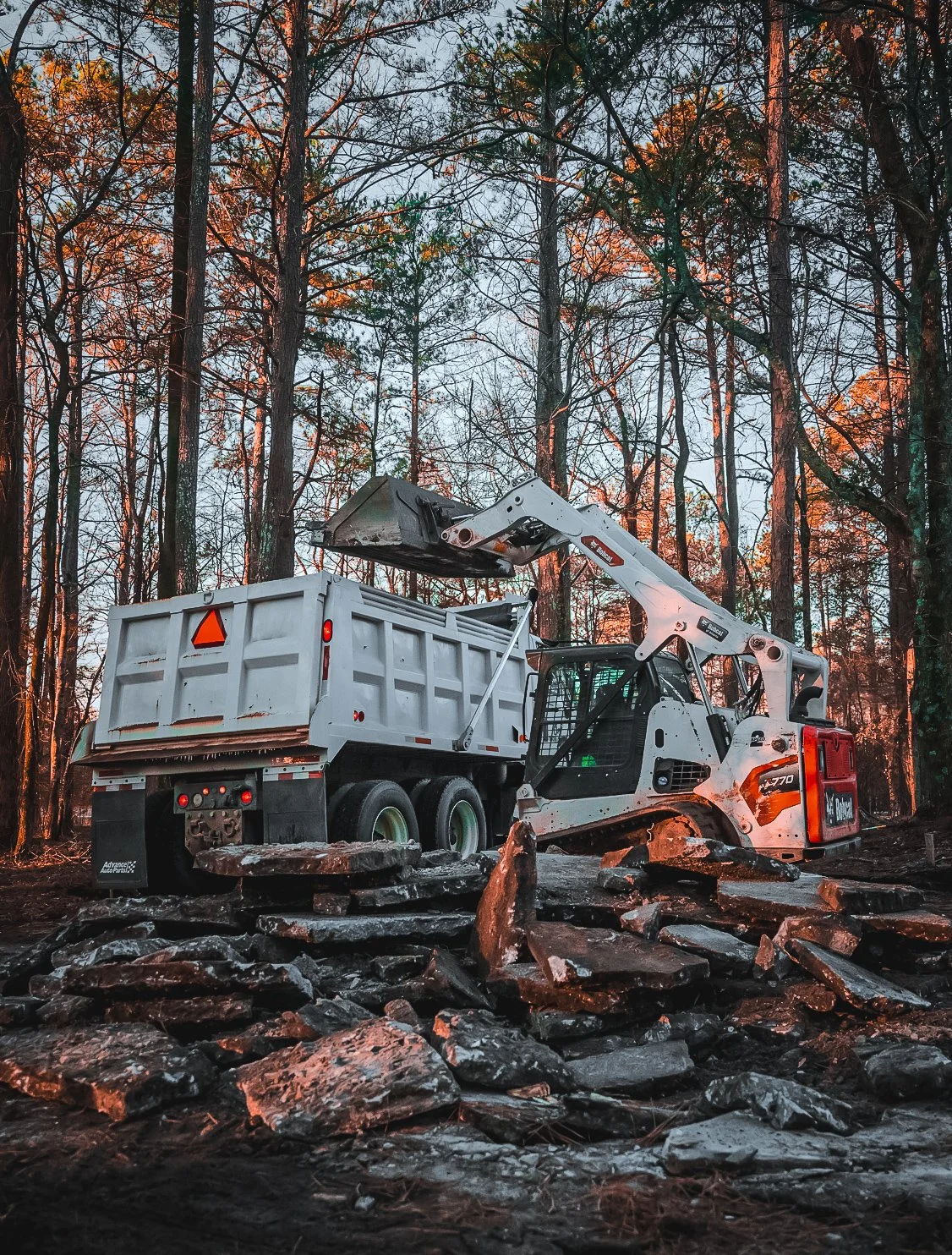 A bulldozer loading rocks into a truck in a forest during sunset.