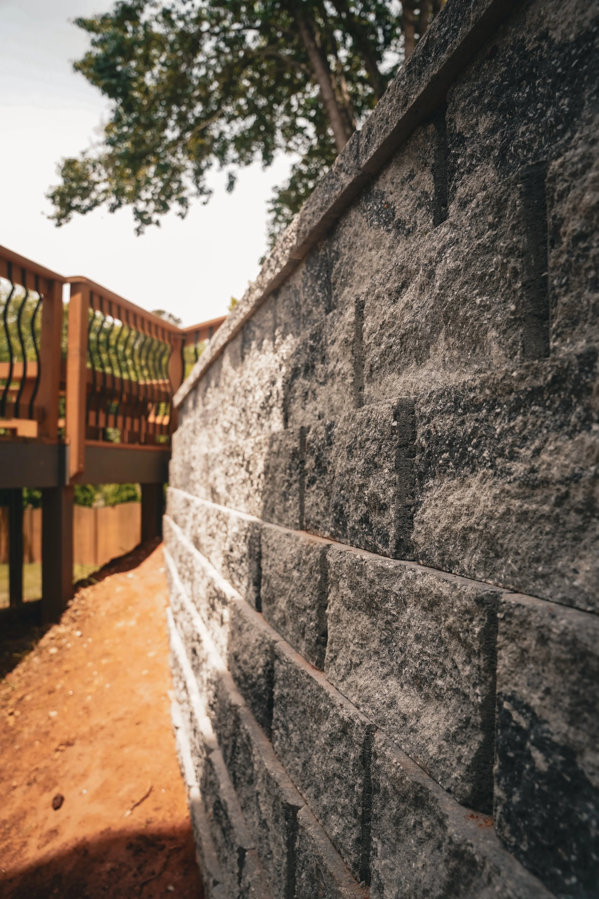 Close-up of a stone brick wall with a wooden deck and railing in the background, trees and sky visible.