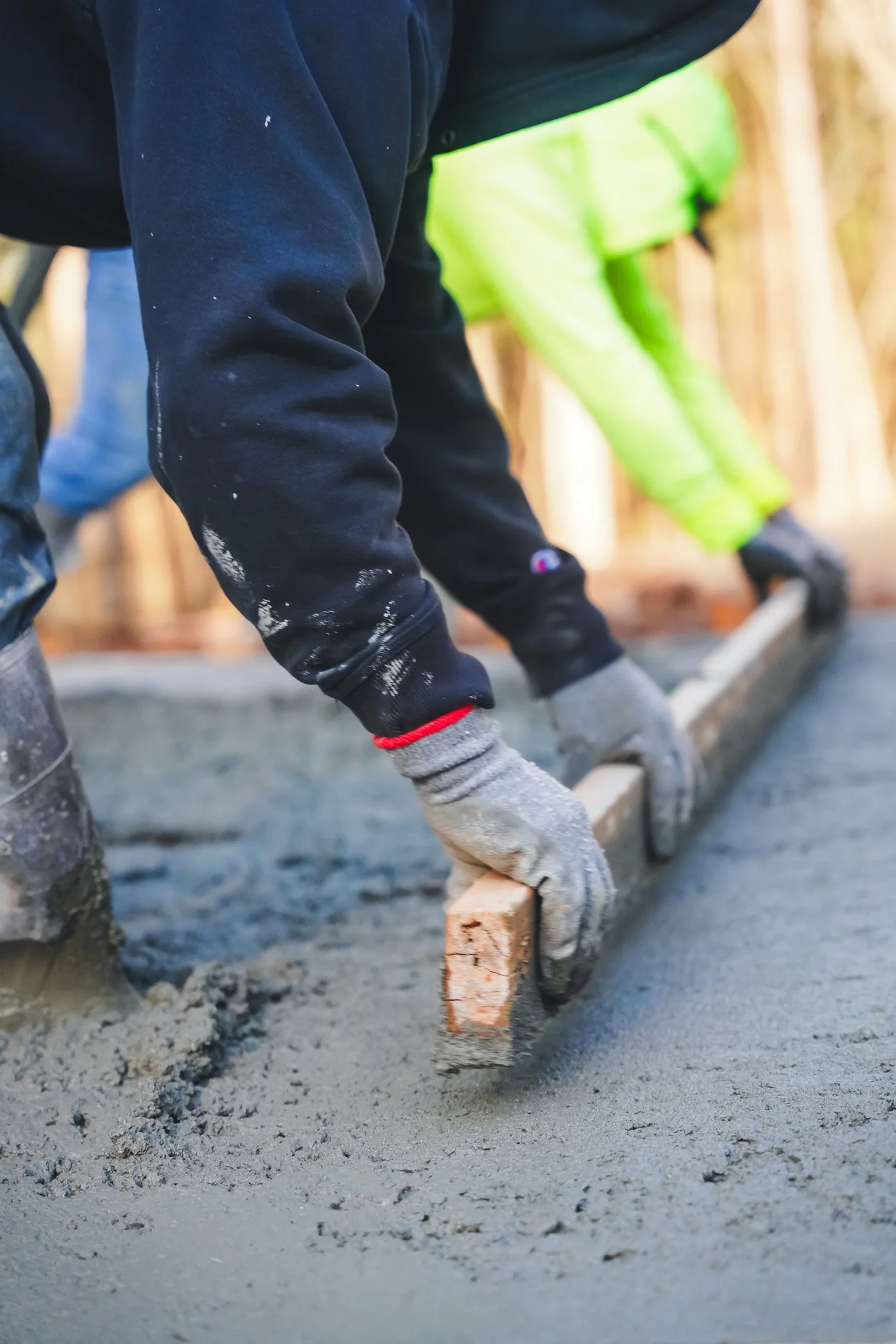 Construction worker smoothing wet concrete with a wooden float trowel, wearing gloves and a black jacket, with another worker in neon green pants in the background.