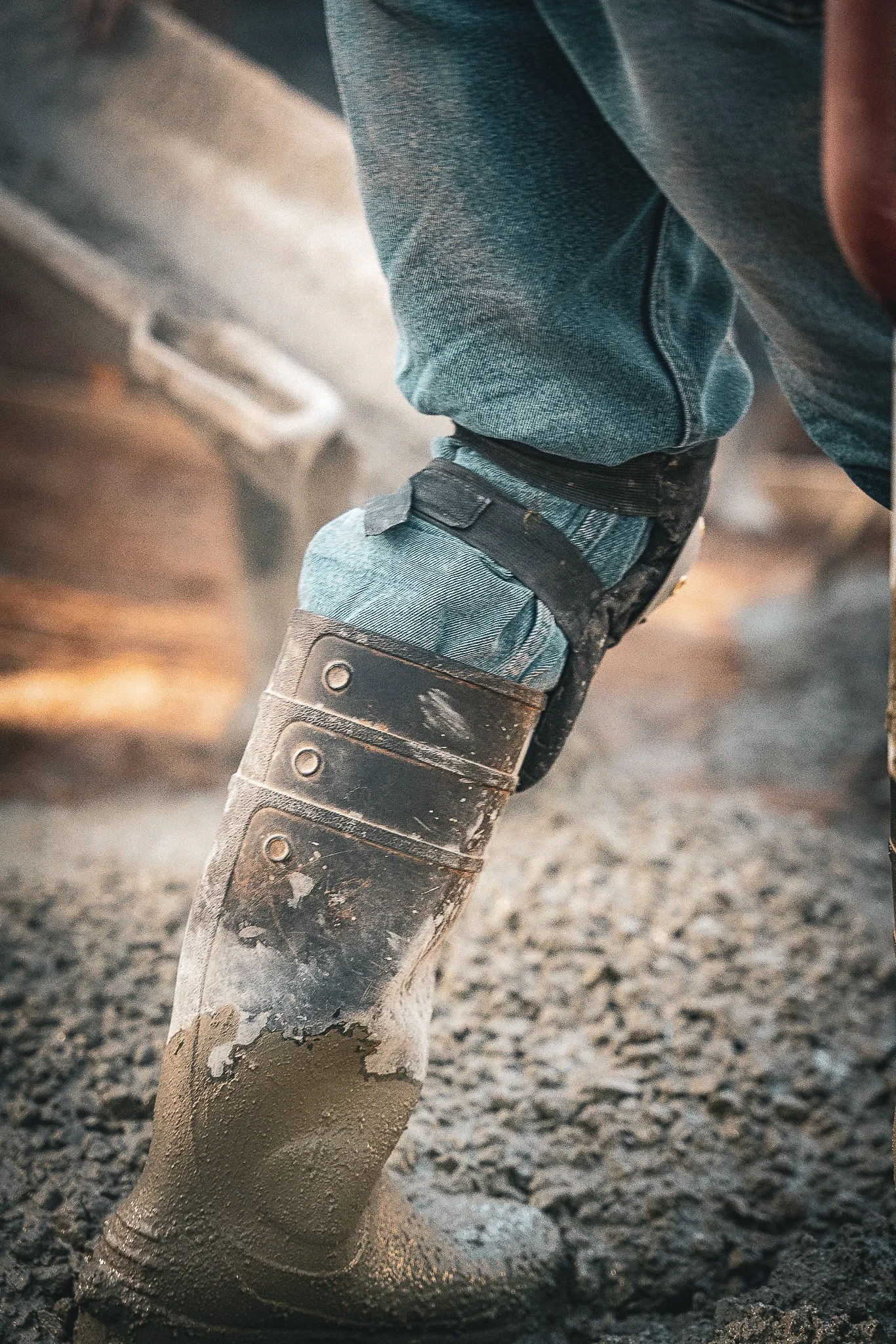 Close-up of a muddy work boot on a gravelly surface, with blue jeans and work pants worn and splattered with mud, in a construction or working environment.