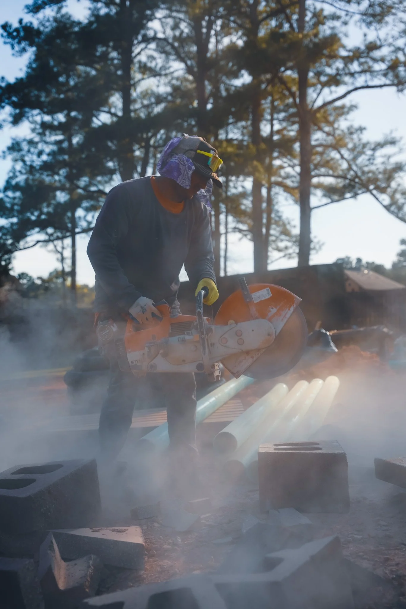 Construction worker using a concrete saw to cut pipes outdoors with trees in the background.