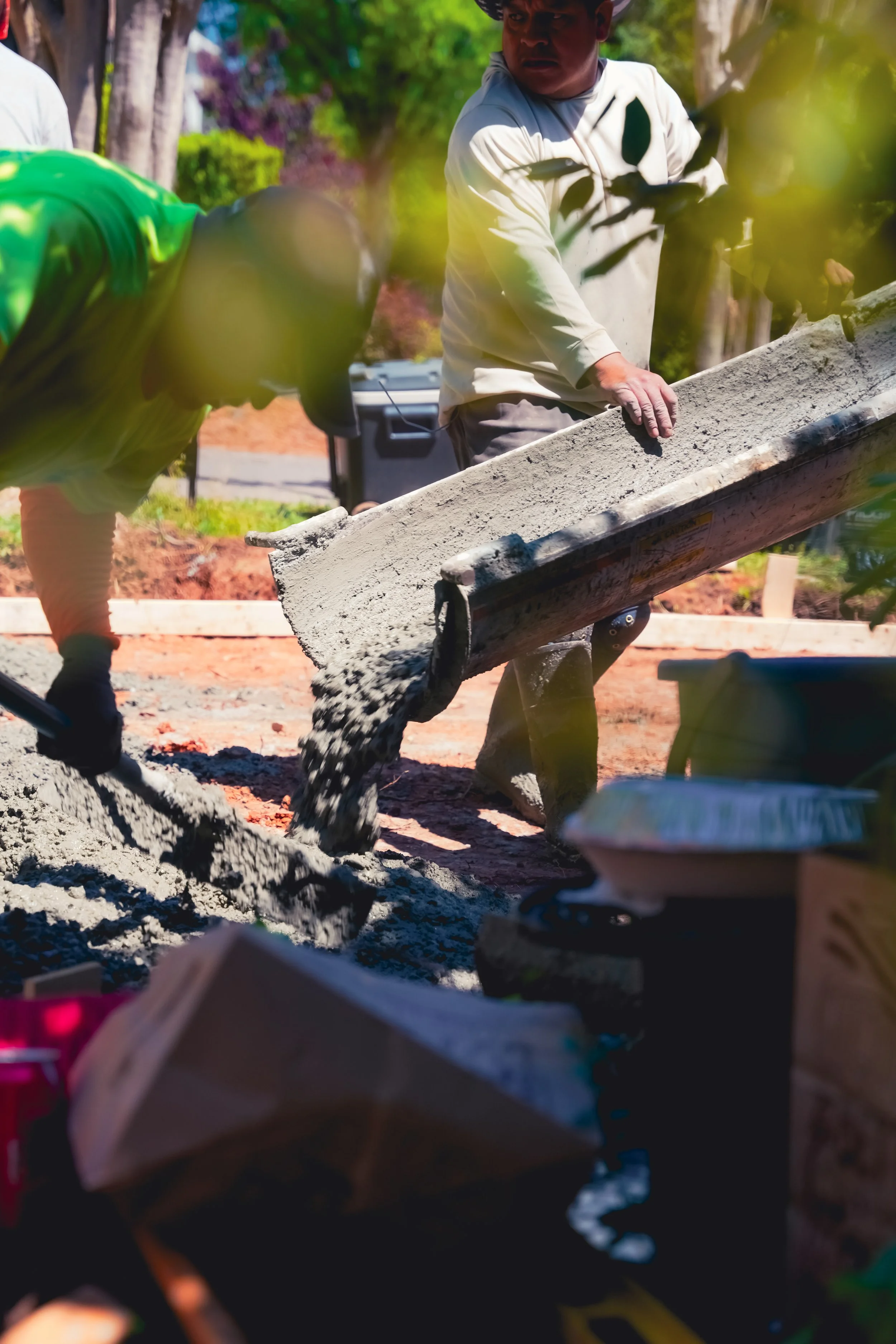 Two men working on pouring concrete in an outdoor setting with trees and plants around.
