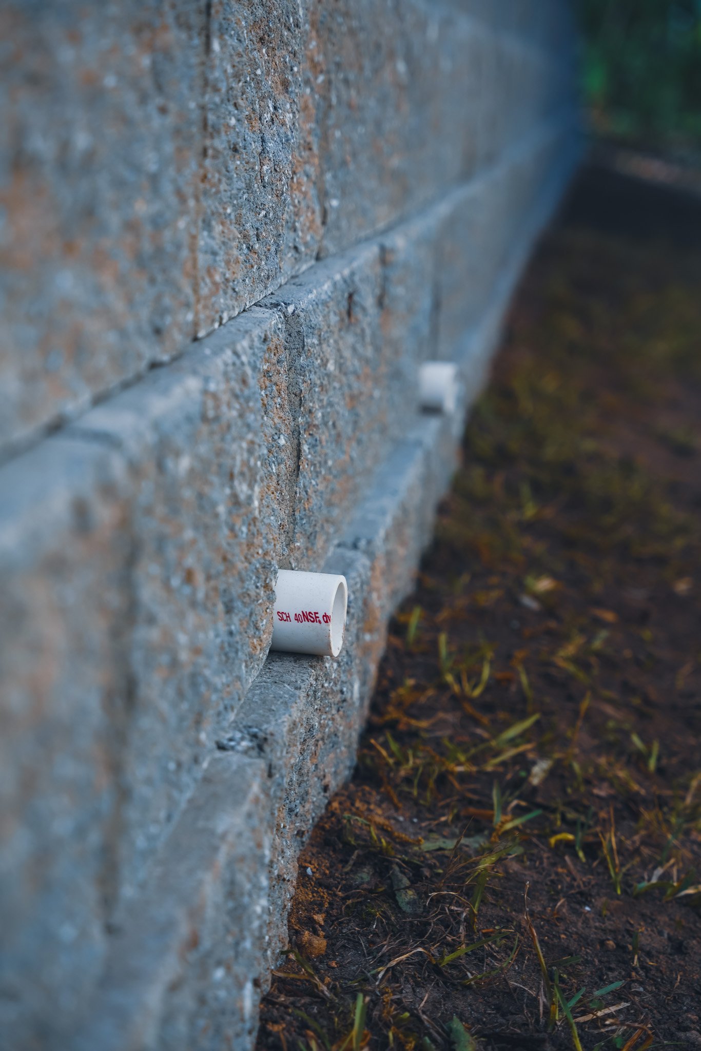 Close-up of a gray brick wall with two small white PVC pipes sticking out near the ground, with some grass growing in the soil beside the wall.