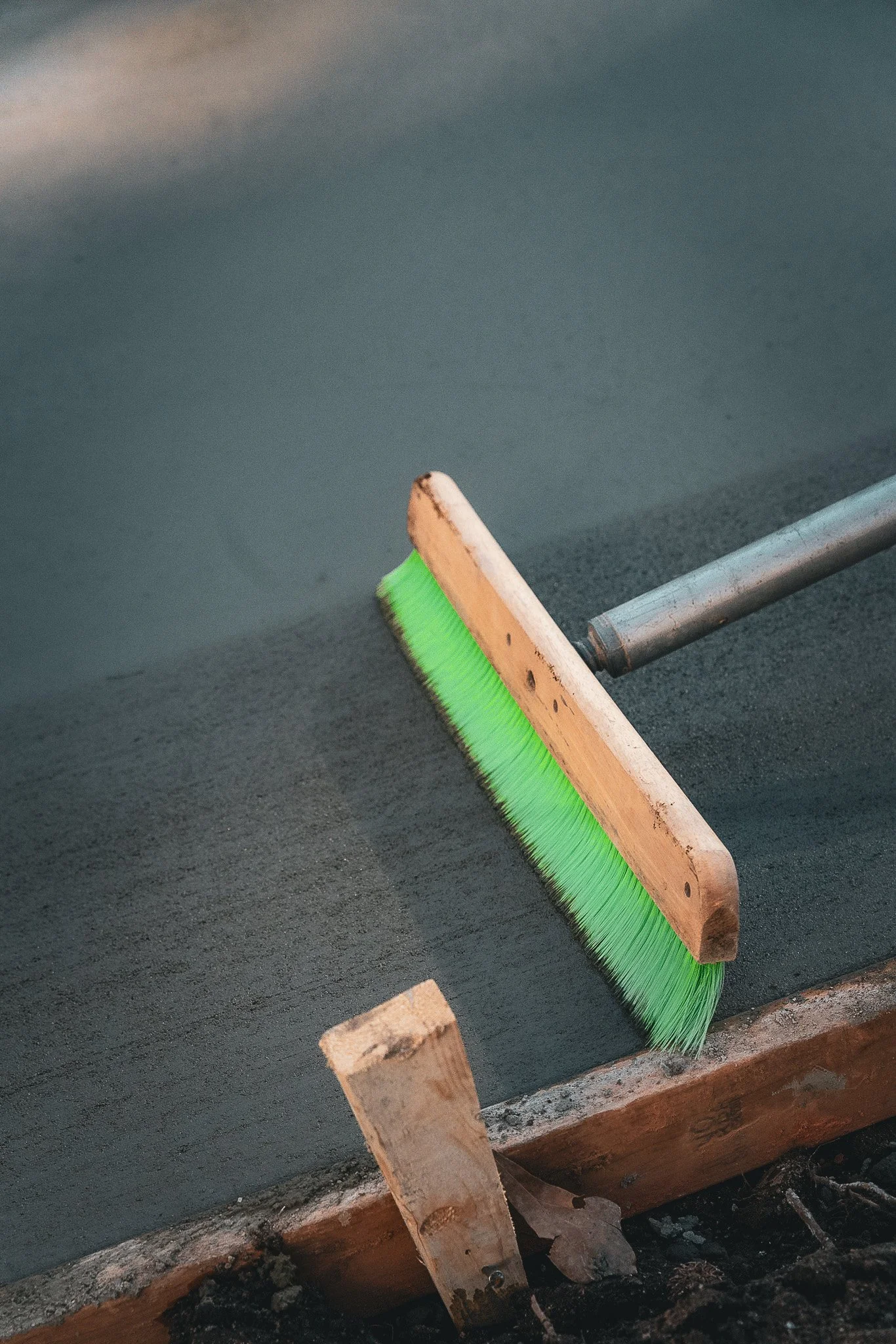 A green broom with a wooden handle sweeping a freshly laid concrete surface.