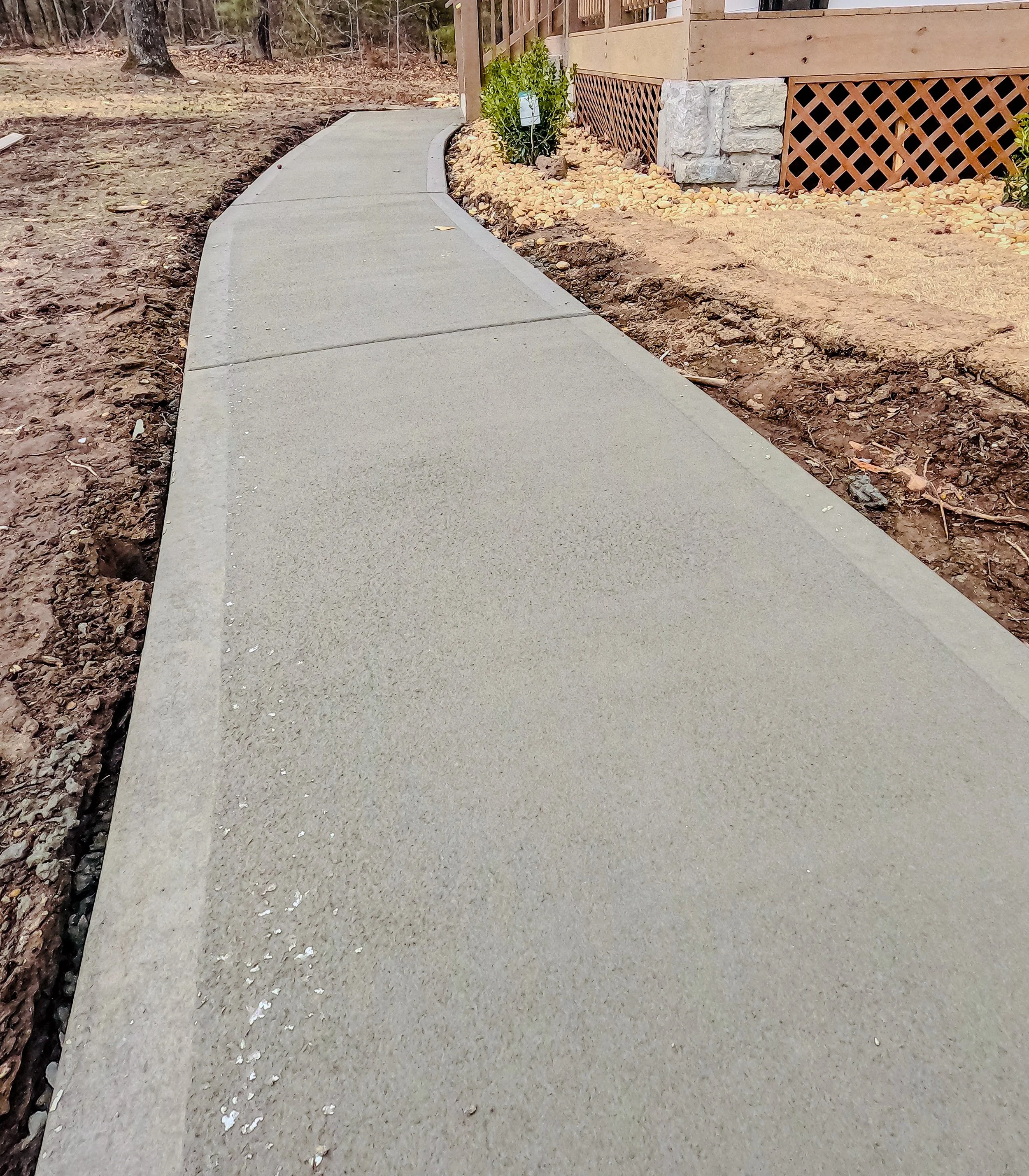 Newly paved concrete sidewalk curving beside a house with a wooden deck and a small plant in the yard.