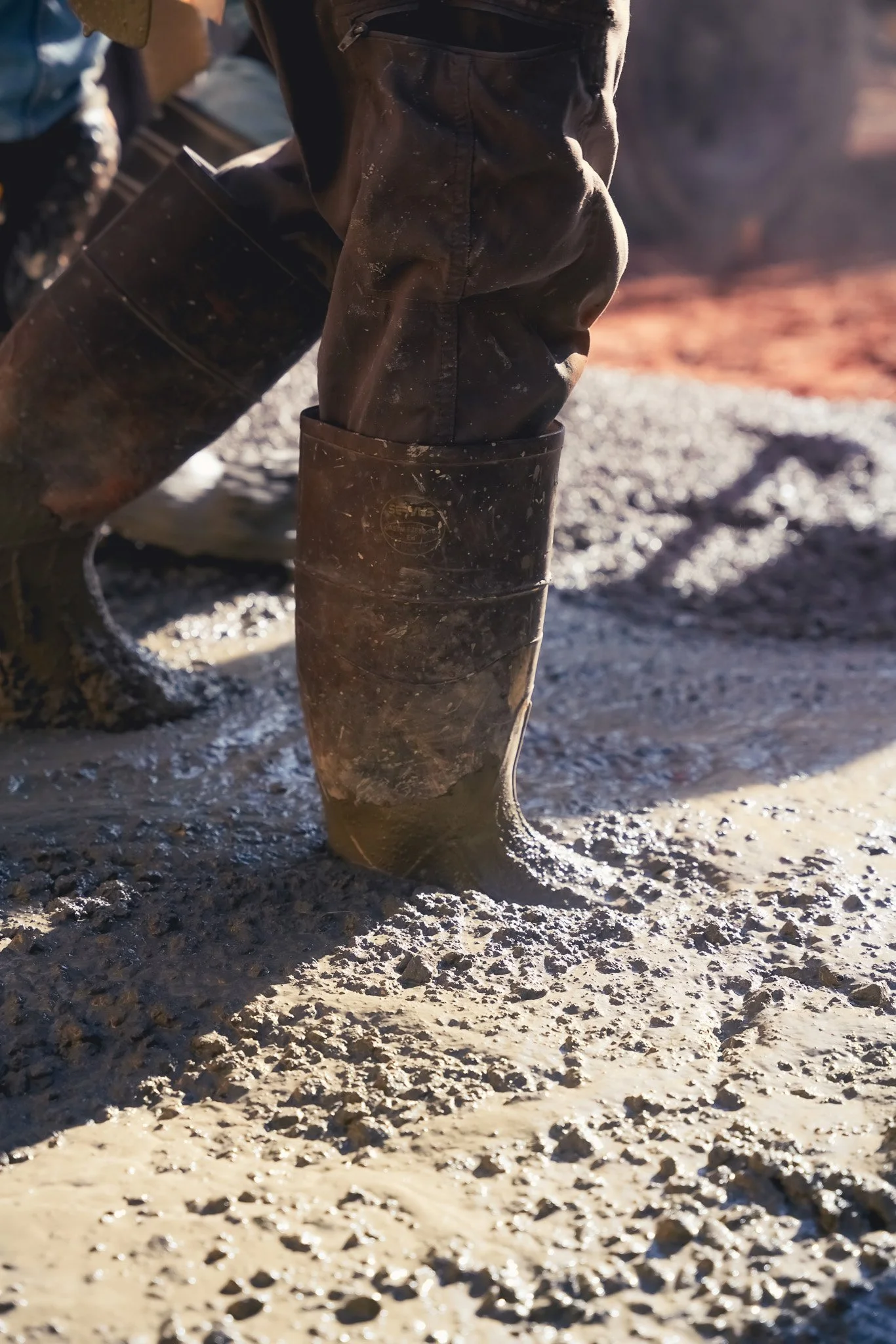 Person wearing mud-covered boots standing on muddy ground, holding another muddy boot in hand.
