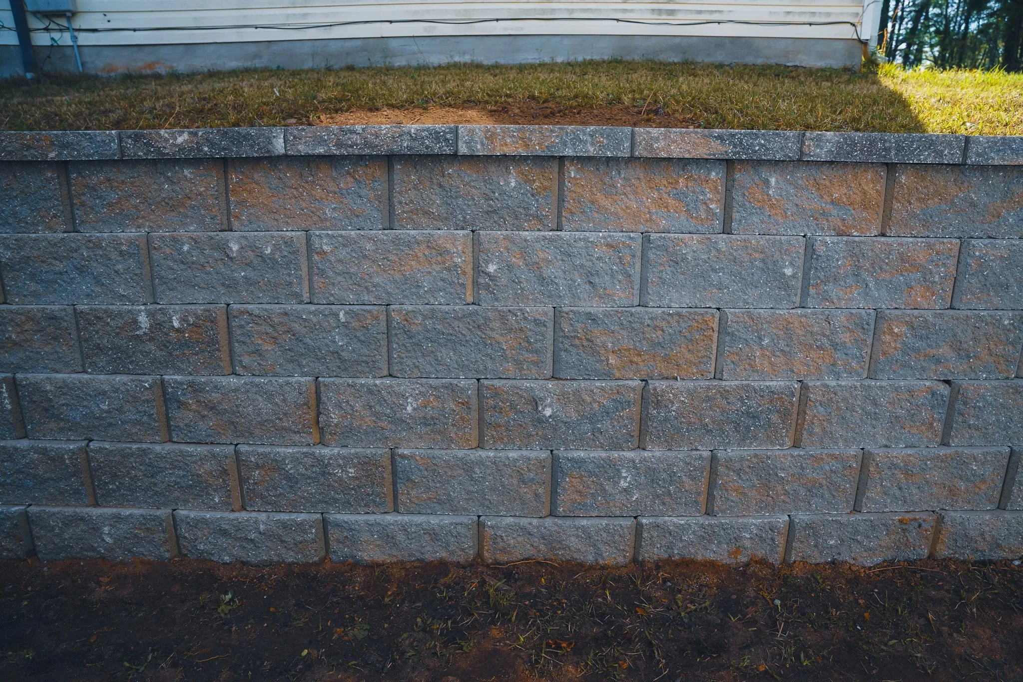 Close-up of a gray brick retaining wall with soil and grass above, in front of a house with white siding.
