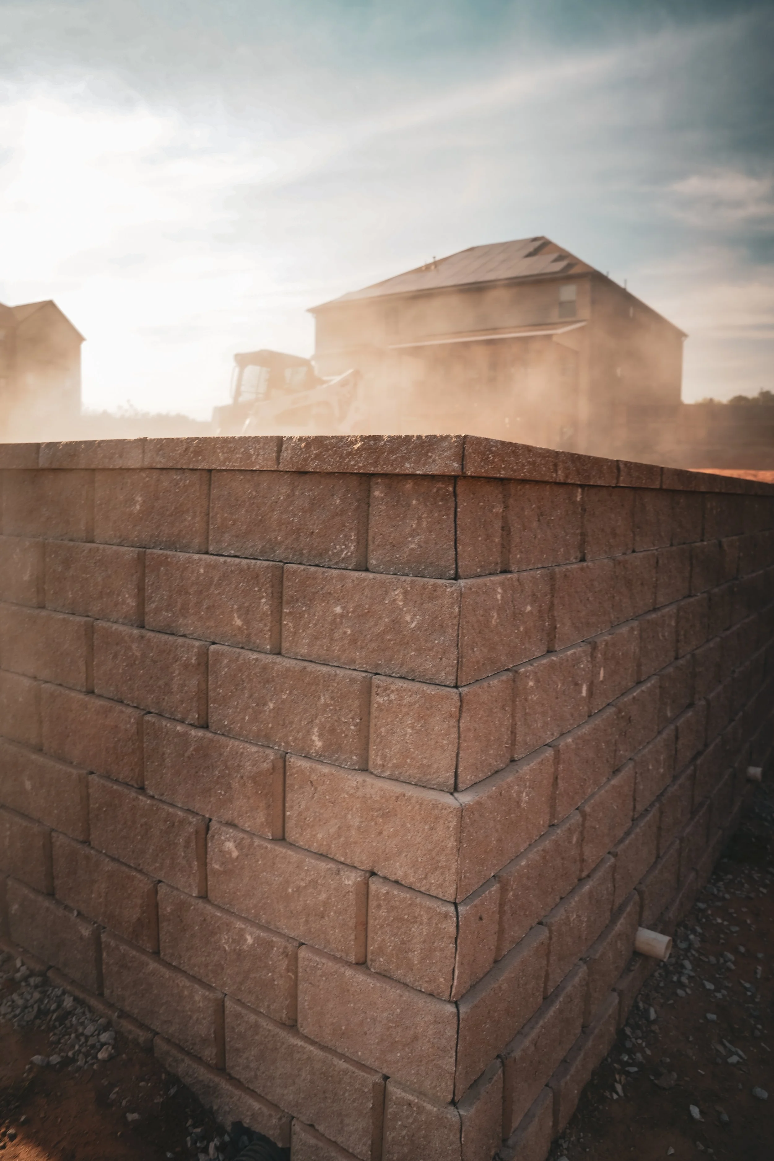 Close-up of a brick wall under construction with house and construction machinery in the background, dust and sunlight in the sky.