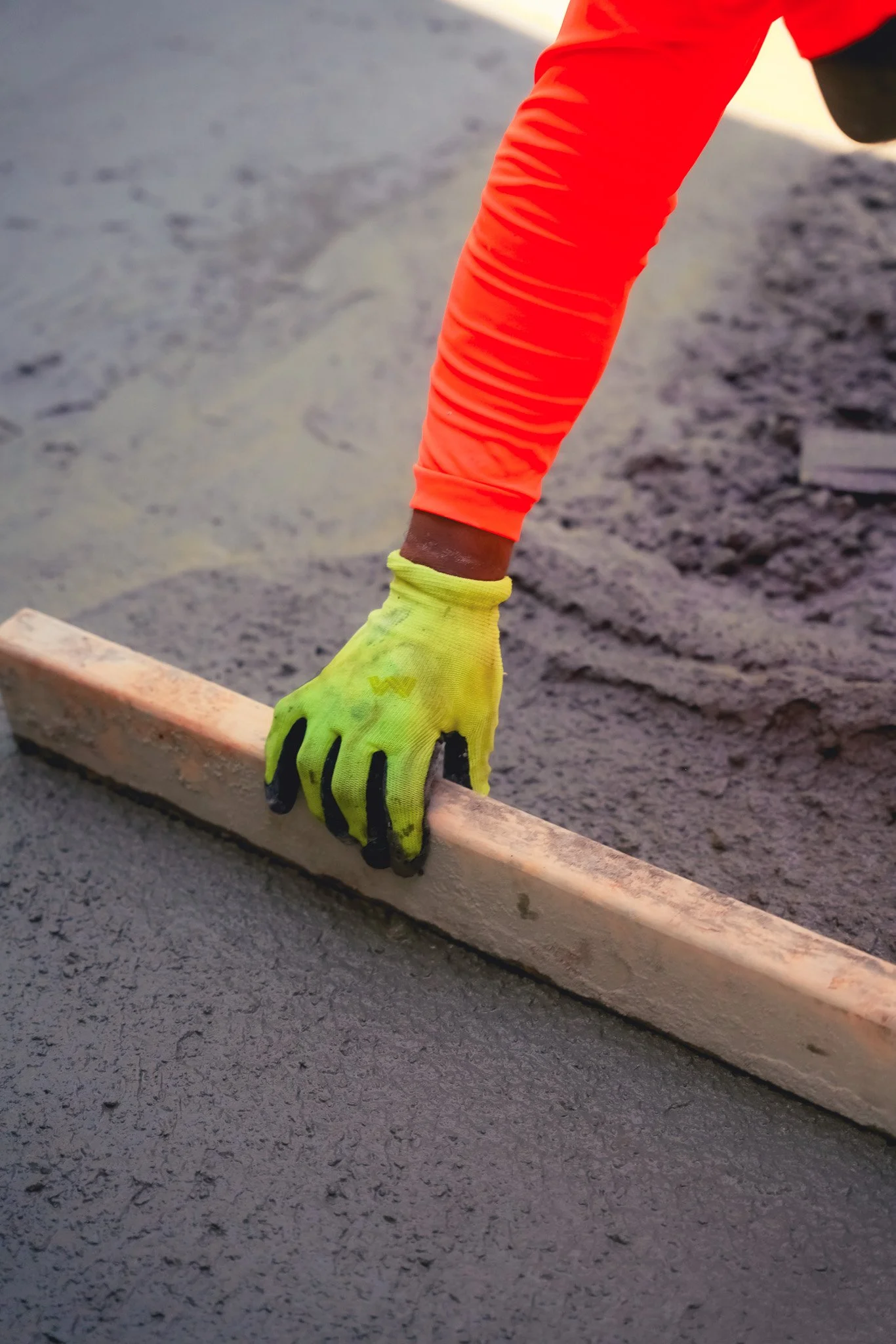 Close-up of a construction worker's hand, wearing a yellow glove, spreading fresh concrete with a wooden float on a surface, with the worker wearing an orange long sleeve shirt.