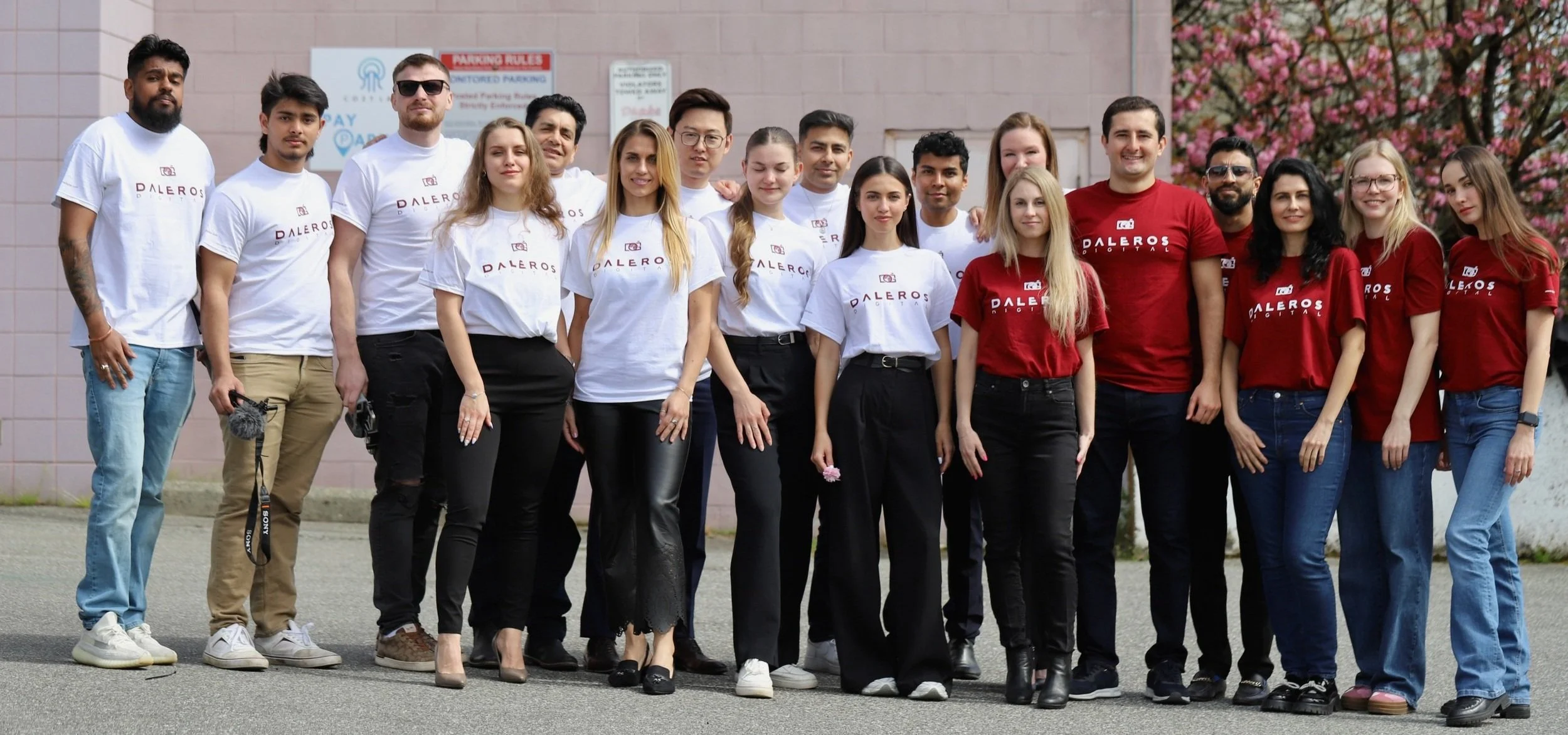 Group of young adults standing outdoors in front of a pink building with a flowering tree, some wearing white T-shirts and others red T-shirts with the logo and name "Daleros".