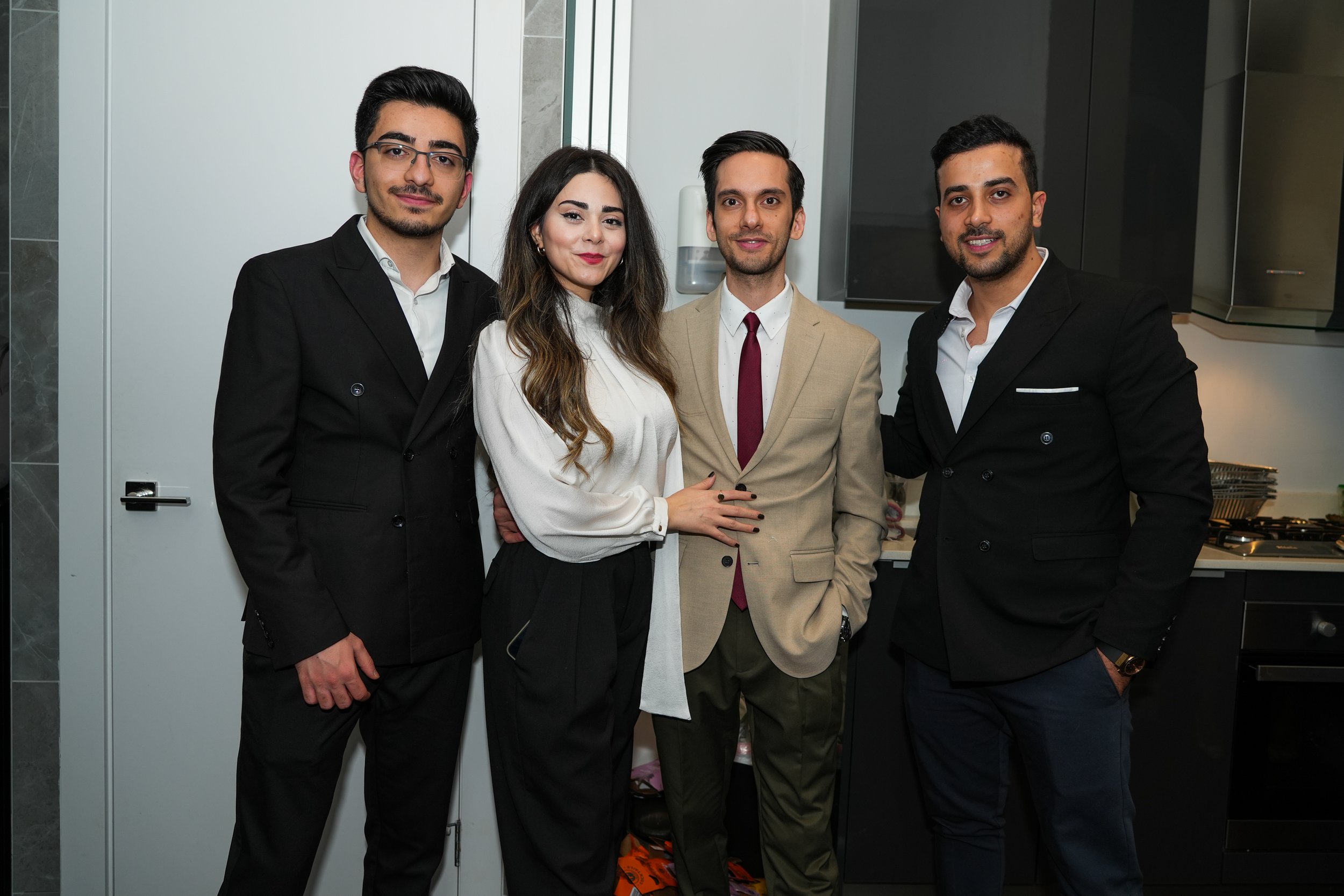 Four people in formal attire posing indoors, with three men in suits and one woman in a blouse.