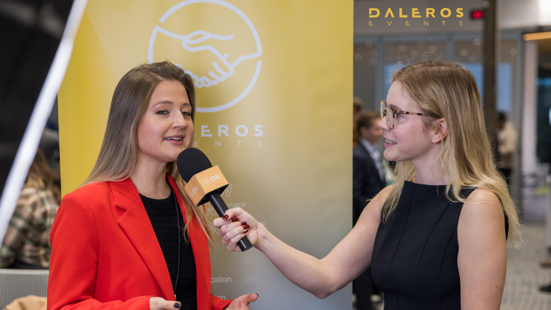 Interviewer with glasses holding a microphone interviewing a woman in a red blazer at an event with a yellow banner in the background.