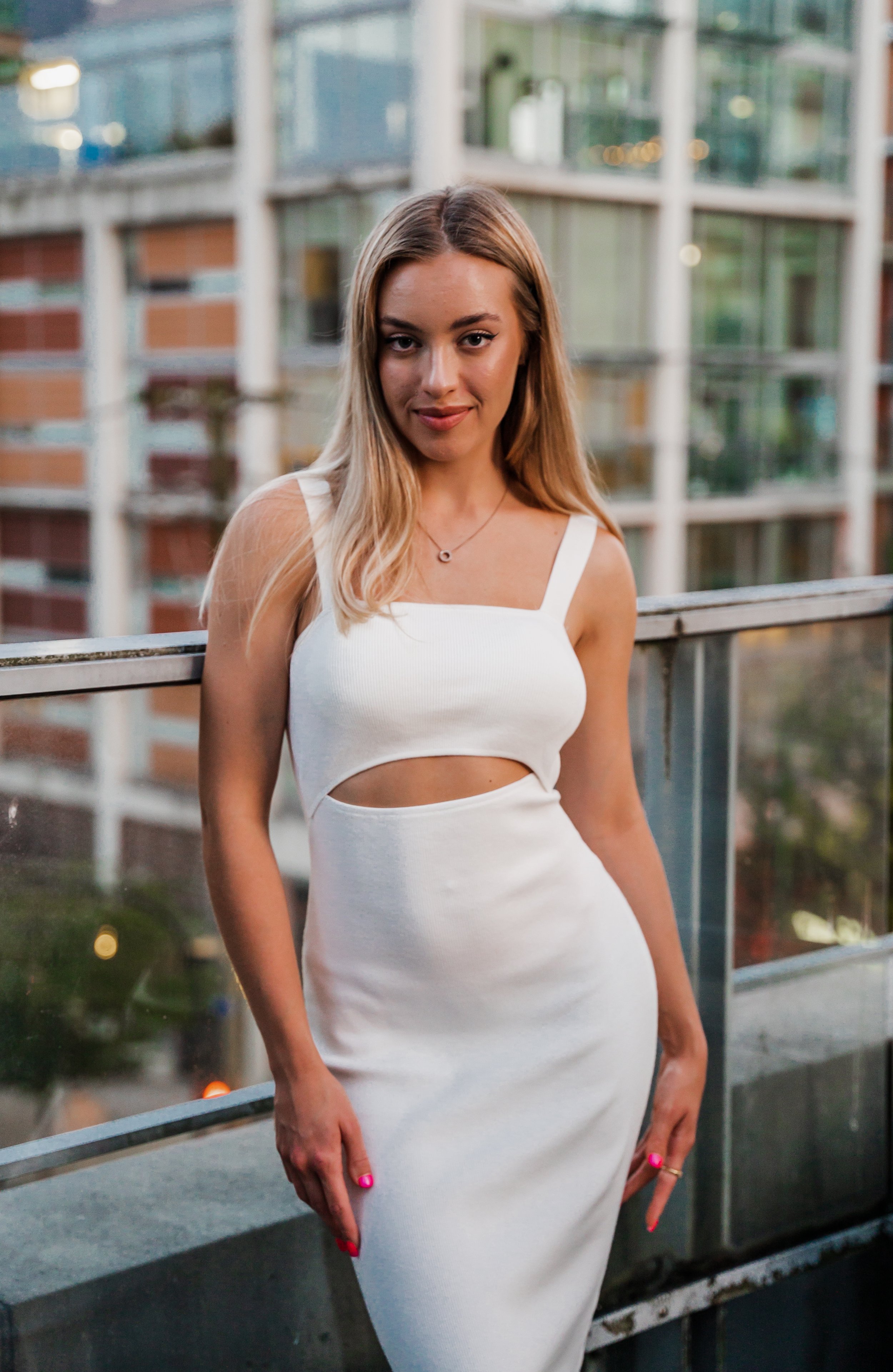 Woman in a white dress standing on a balcony with an urban background.