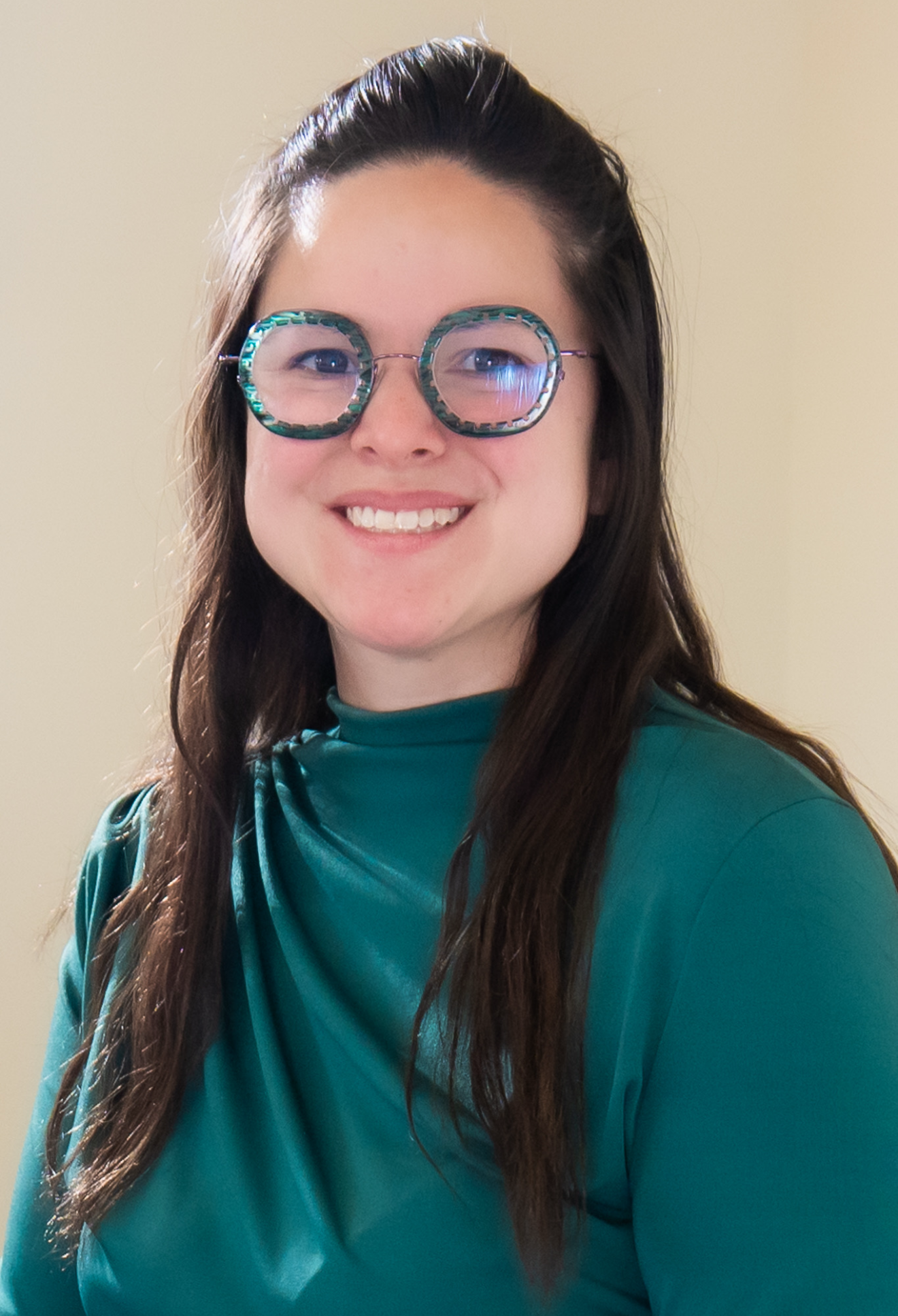 Close-up of a smiling woman with long brown hair wearing large, patterned glasses and a teal top with a draped collar.