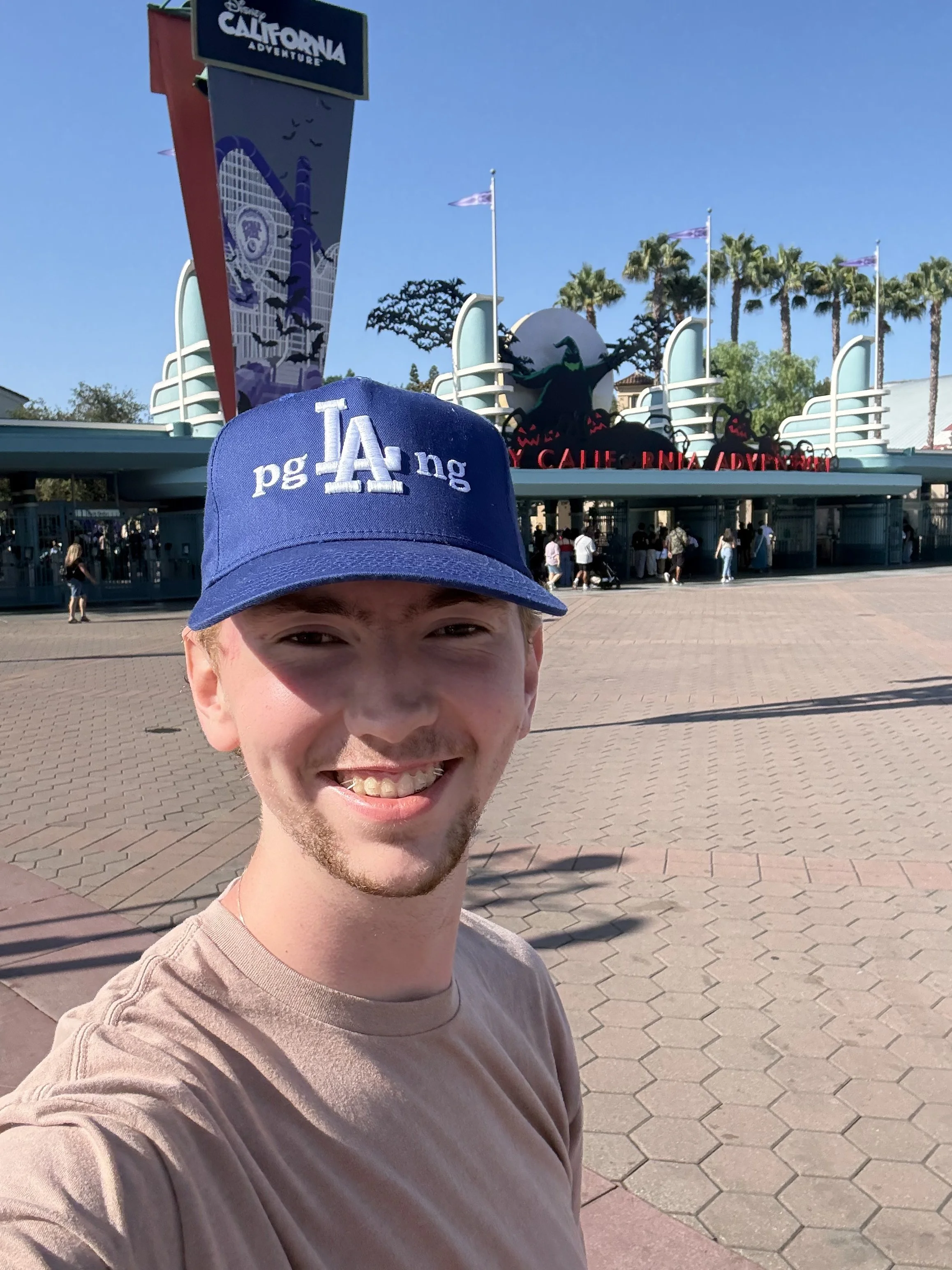 A smiling man wearing a blue Los Angeles Dodgers cap taking a selfie in front of California Adventure theme park entrance with a blue sky overhead and some palm trees in the background