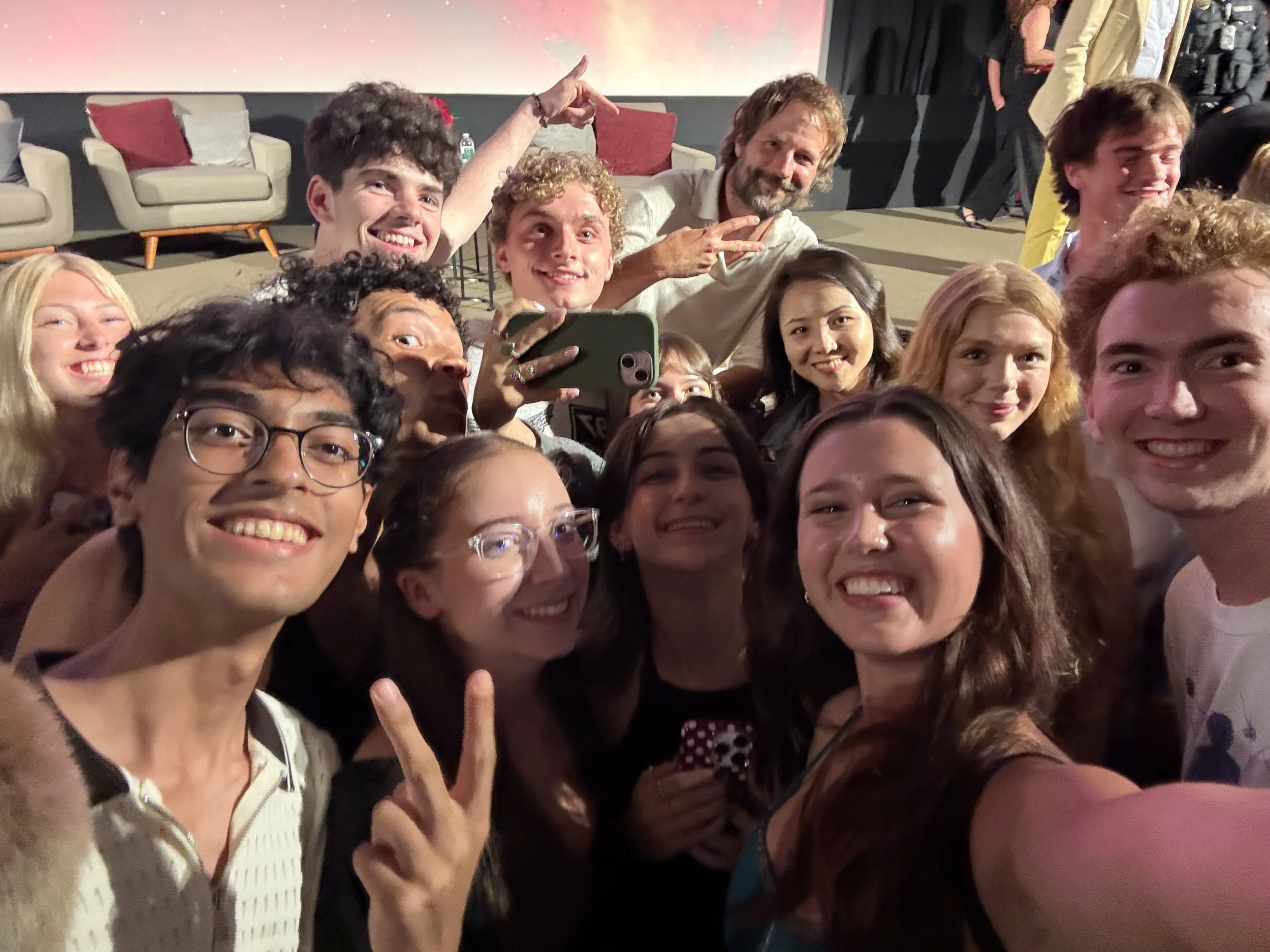 Group of smiling people taking a selfie at an event, with couches and a colorful wall in the background.