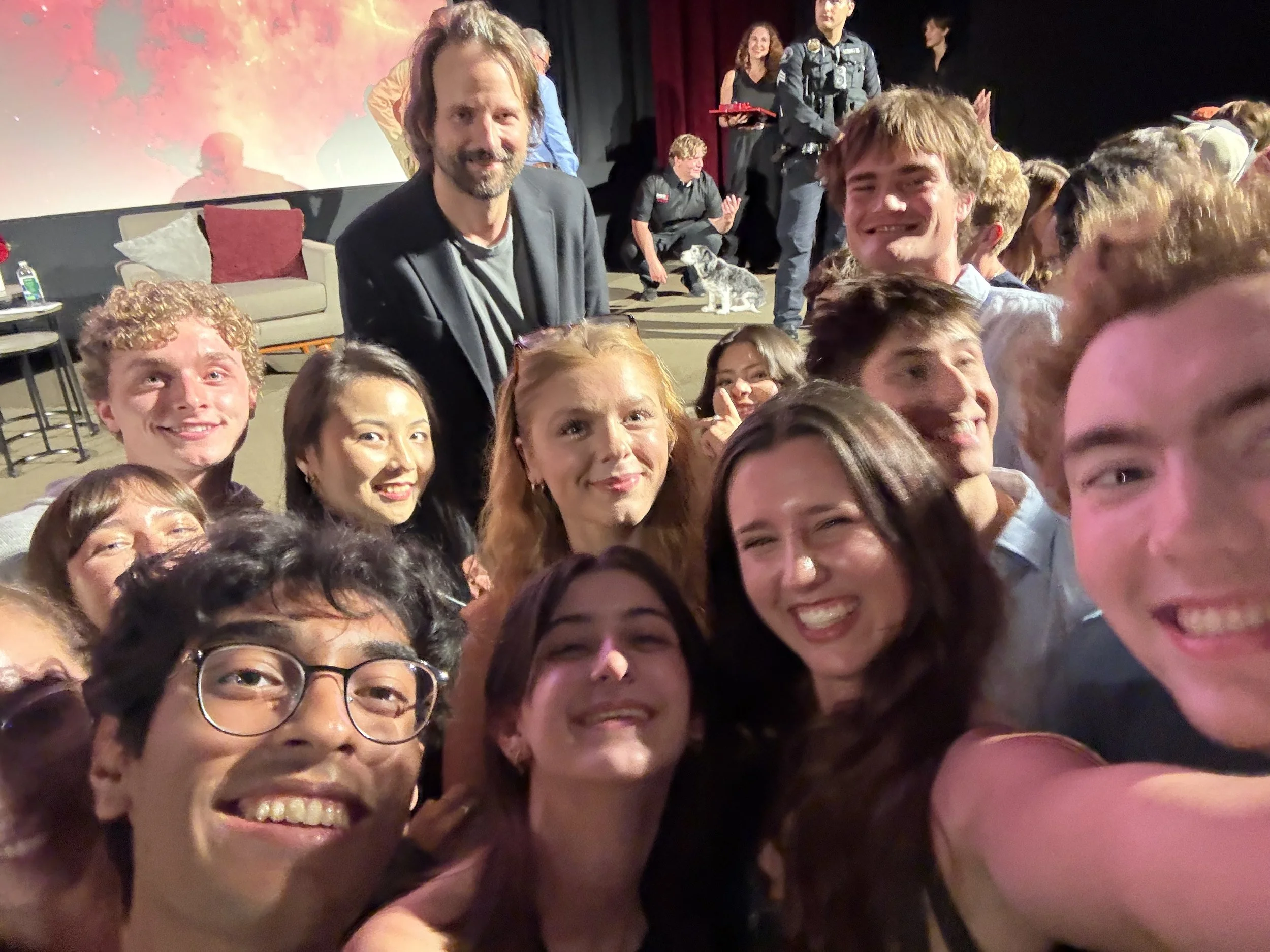 A group of people taking a selfie at an indoor event, with some seated and some standing, including a man with long hair and a beard in the background and a woman with curly red hair.