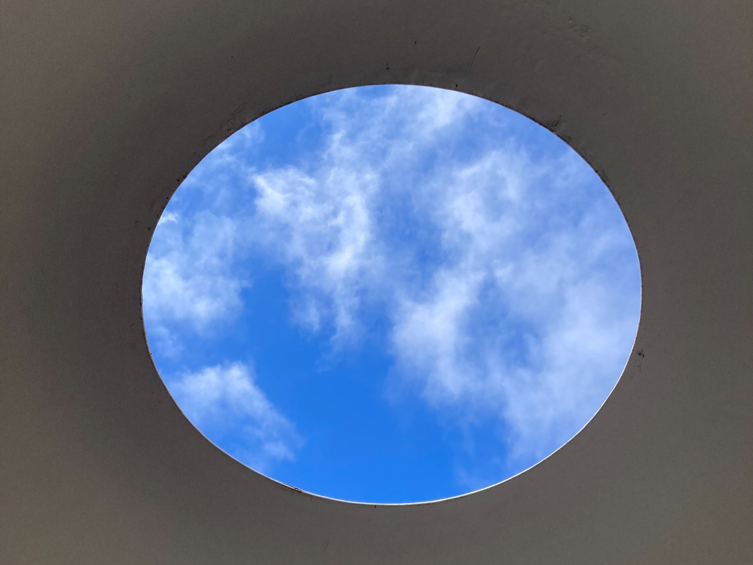 Blue sky with some scattered white clouds viewed through a circular opening in a gray ceiling.