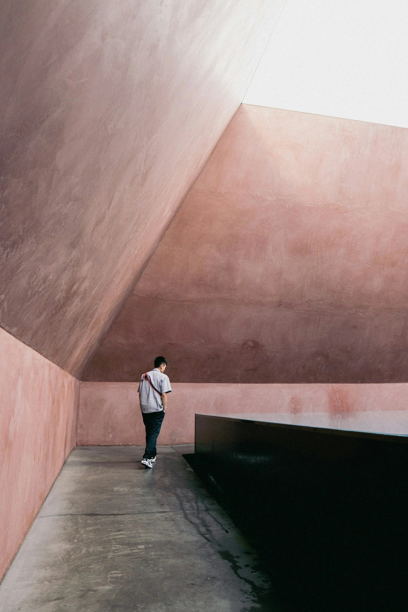 A person walking beside a reflecting pool in a minimalist, pink-hued modern architectural space with geometric walls and ceiling.