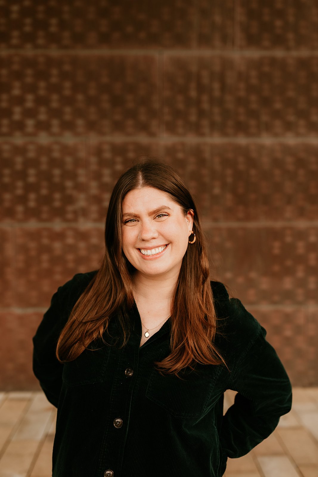 A young woman with long brown hair, smiling, wearing a black jacket, standing against a brick wall background.