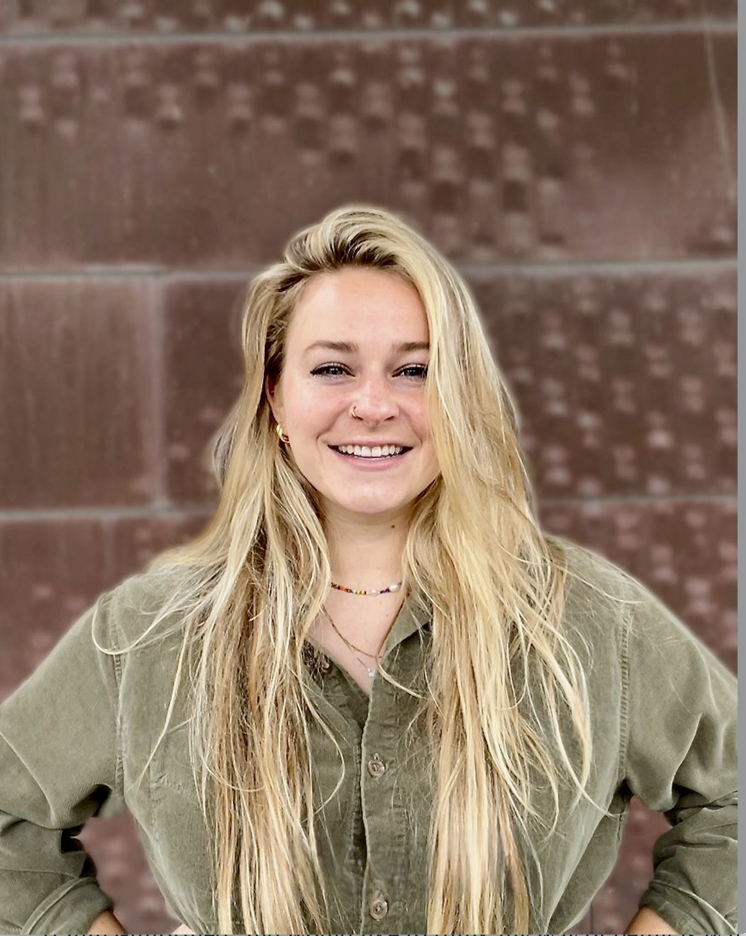 A young woman with long blonde hair, smiling, wearing an olive green button-up shirt and jewelry, standing against a red brick wall background.