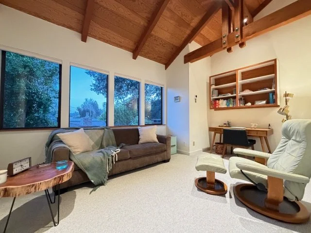 Living room with a brown sofa, white armchair, wooden ceiling with beams, large windows showing trees outside, and a wooden desk with a black chair.