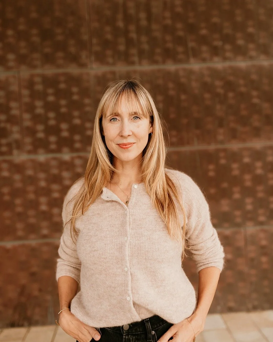 A woman with blonde hair, wearing a light beige cardigan and black pants, standing outdoors in front of a textured brown wall.