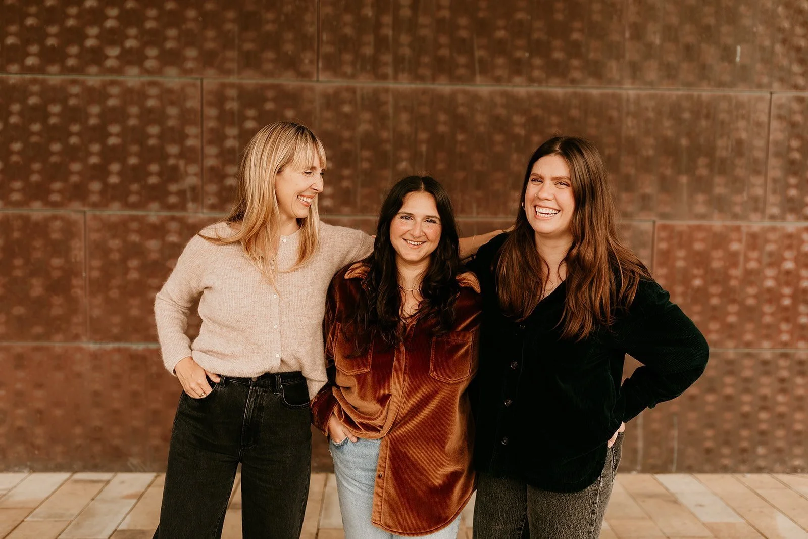 Three women standing together, smiling and laughing, against a textured wall background, with the woman on the right having her arm around the woman in the middle.