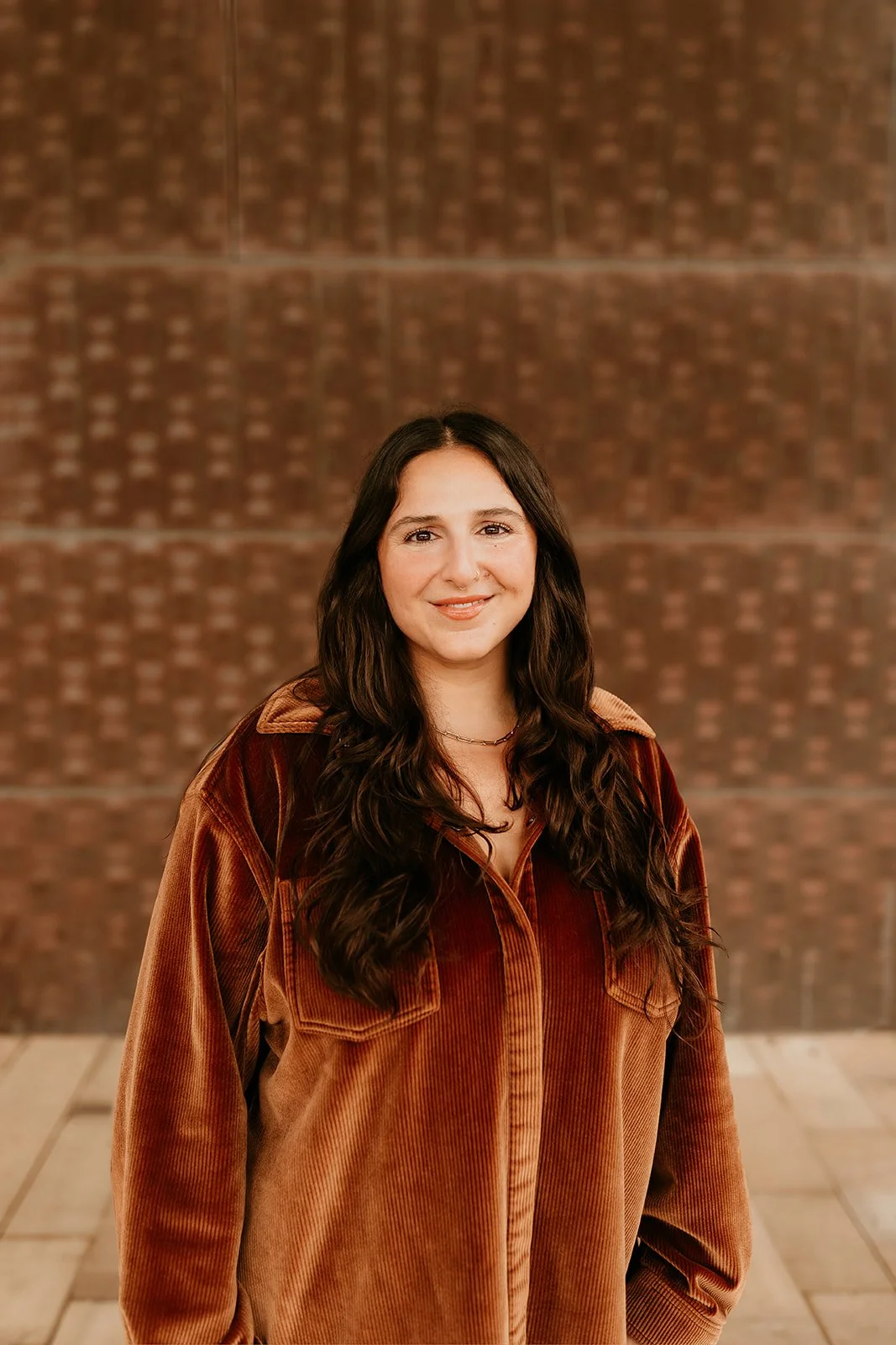 A young woman with long dark hair wearing a brown corduroy jacket and a small necklace, standing in front of a large brick wall, smiling at the camera.