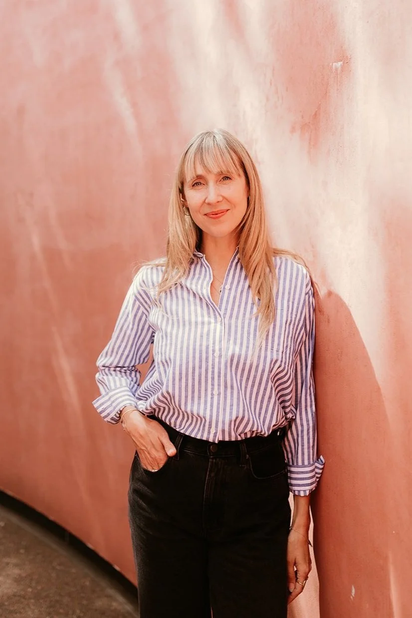 A woman with blonde hair wearing a striped button-up shirt and black pants, standing next to a pink wall.