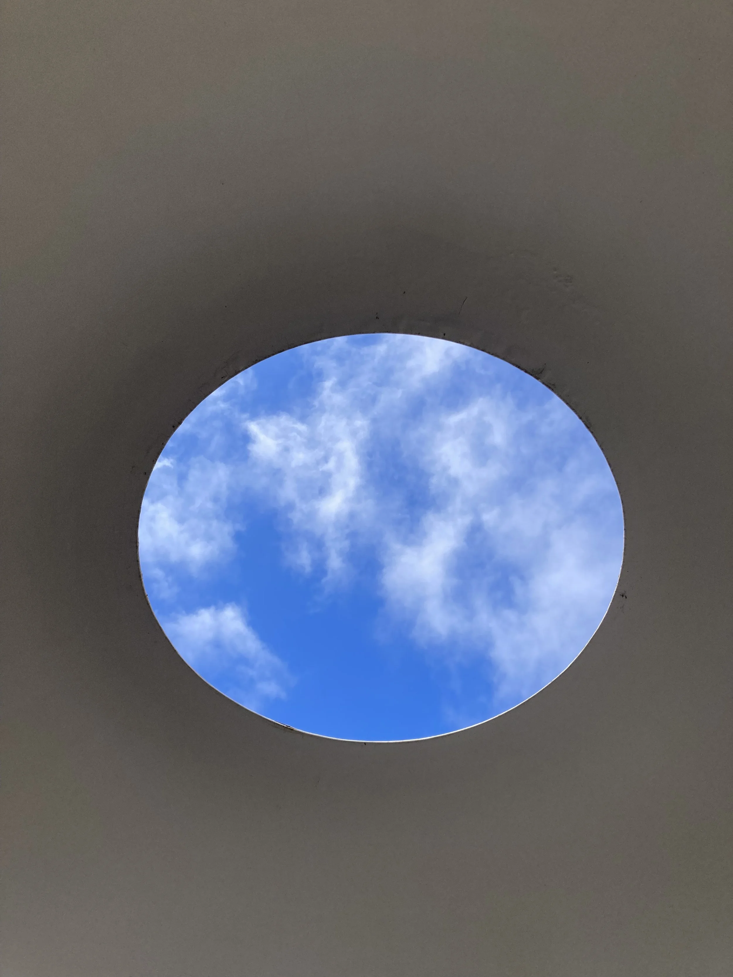 Blue sky with scattered white clouds viewed through a circular opening in a gray ceiling.