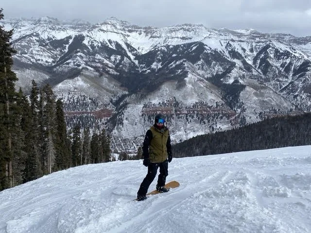 Person wearing a green jacket, black pants, and a blue helmet snowboarding on a snowy slope with a mountain range and pine trees in the background.