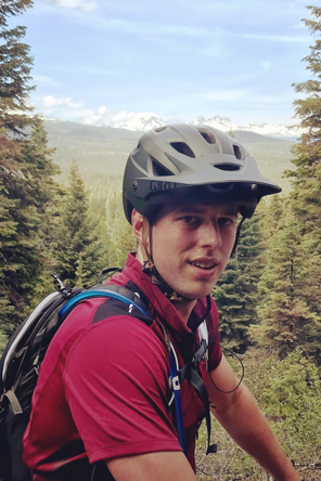 A young man wearing a helmet and red shirt outdoors, with trees and mountains in the background.