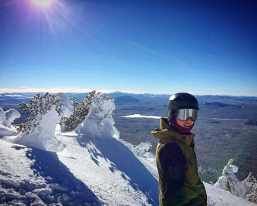 A person wearing a helmet and ski goggles standing on a snowy mountain with snow-covered trees, mountains, and a clear blue sky in the background.