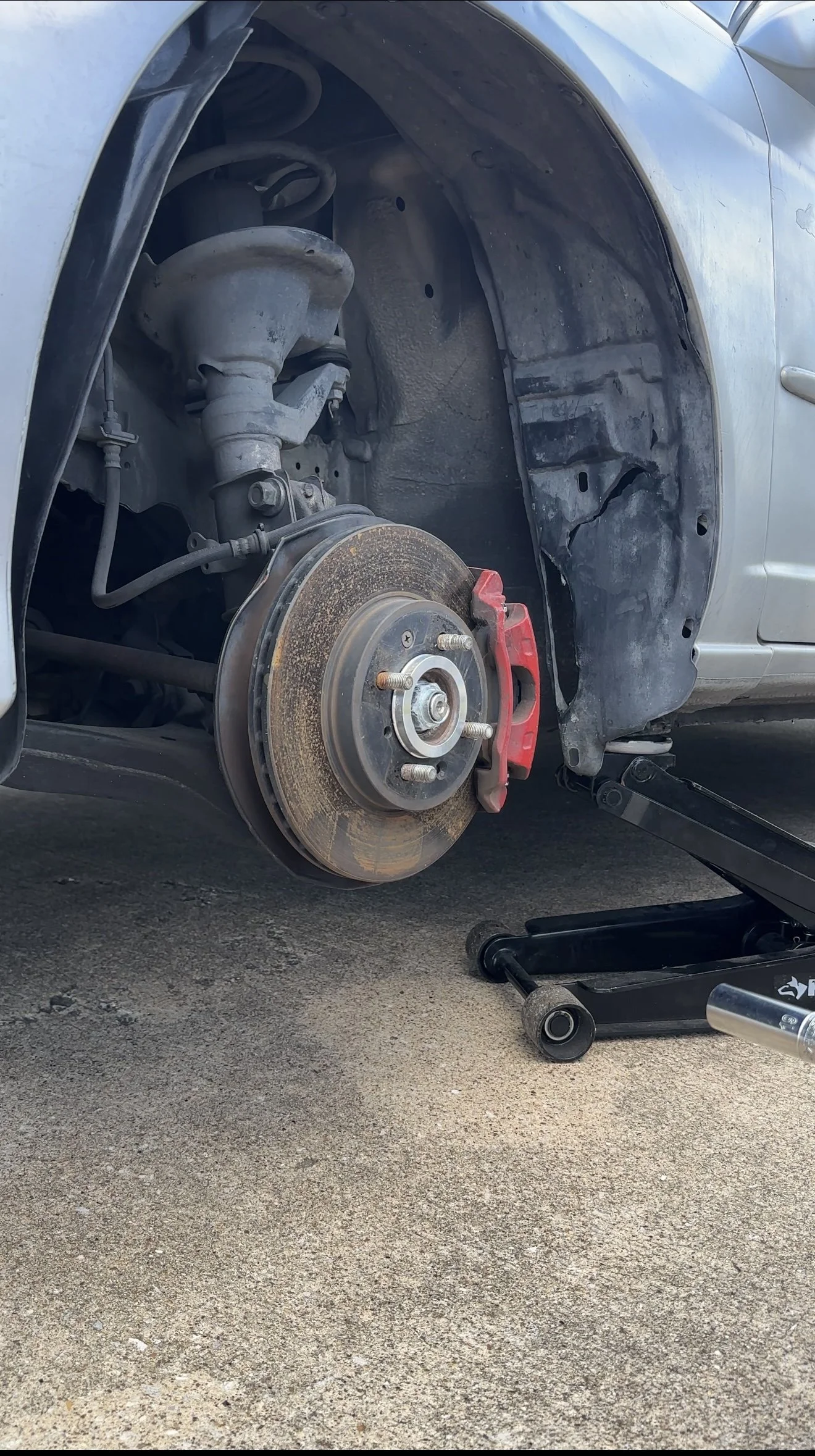 A silver car with the front wheel removed, showing the brake rotor and caliper, on a concrete driveway with a hydraulic floor jack lifting the vehicle.