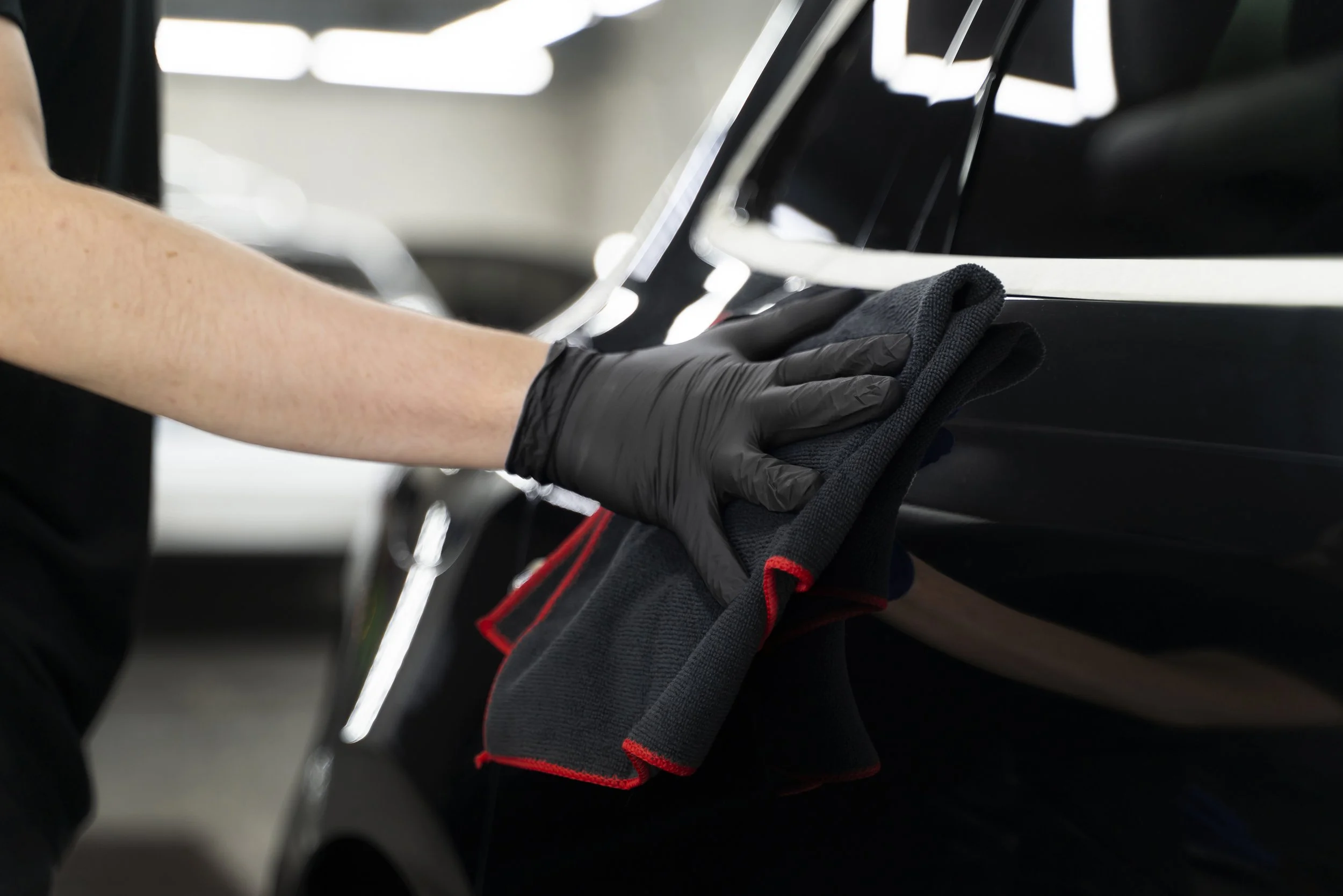 Person wearing black gloves cleaning a black car with a cloth in a garage.