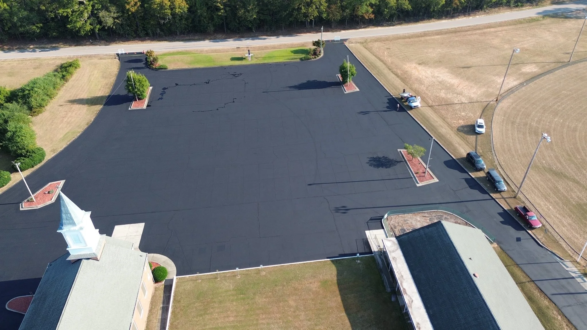 Empty parking lot with a few cars parked along the edge, small trees in landscaped areas, and a building with a steeple on the left.