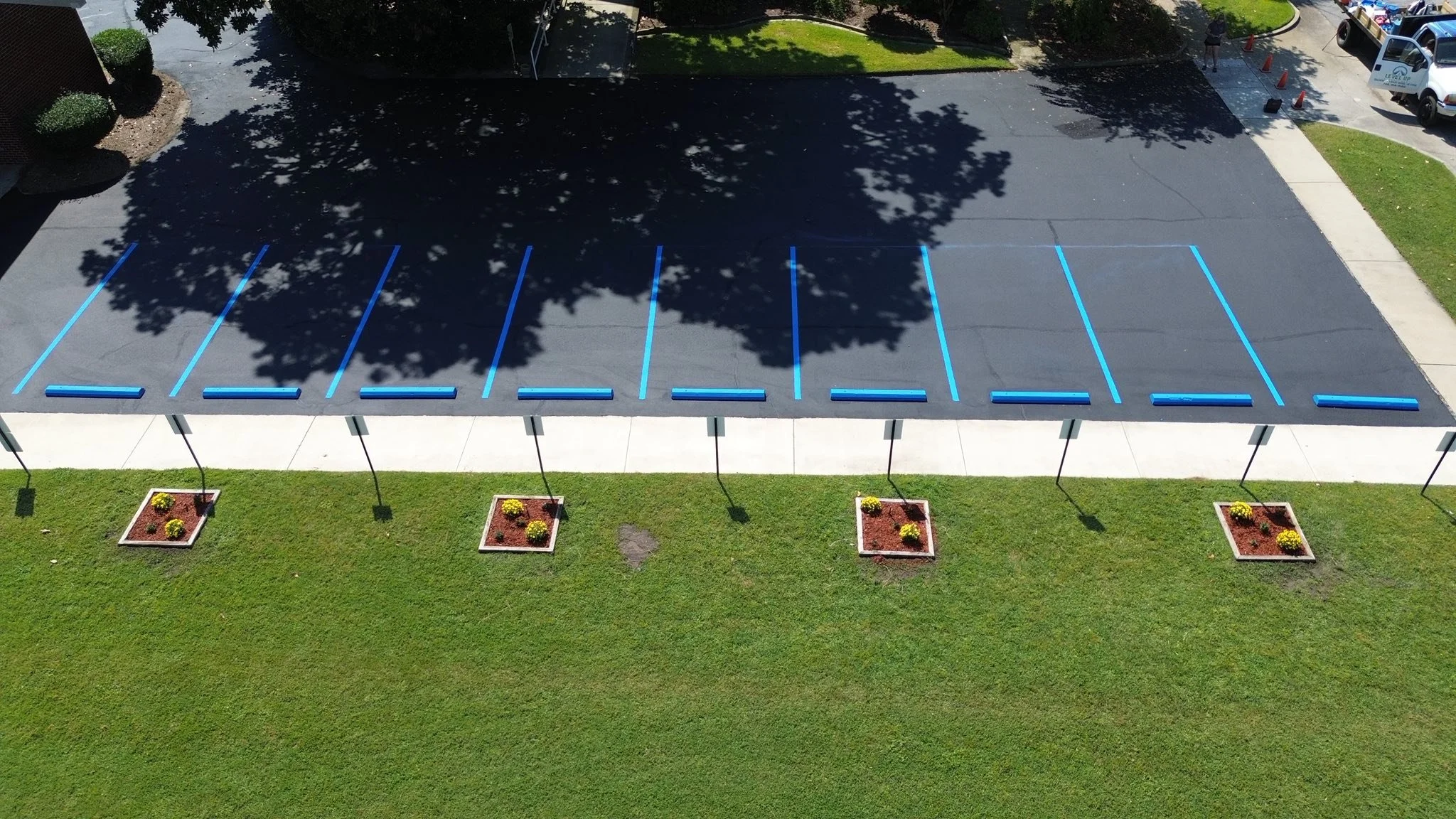 An aerial view of a parking lot with empty parking spaces marked with blue lines, adjacent to a sidewalk and grassy area with small flower beds, and trees casting shadows over part of the lot.