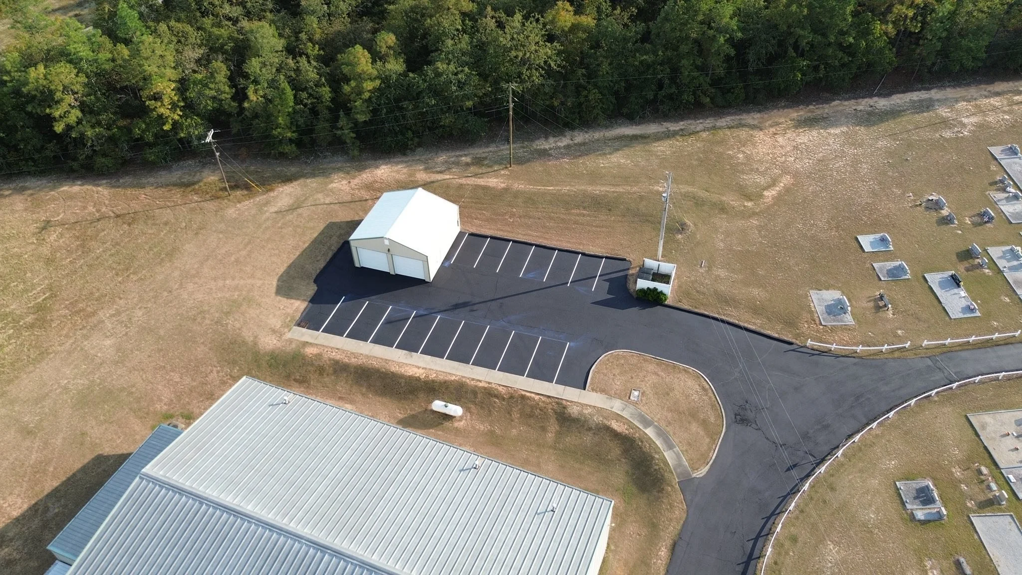 Aerial view of a small parking lot next to a building with a metal roof, and a neighboring area with multiple grave markers in a cemetery. There are trees in the background, utility poles, and a white storage shed in the parking area.