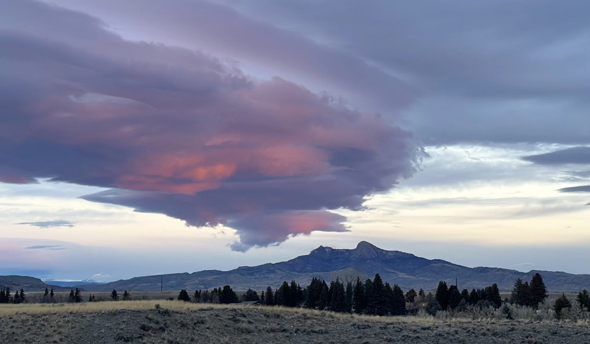 Dramatic sky with colorful clouds over a mountain landscape, with trees in the foreground.