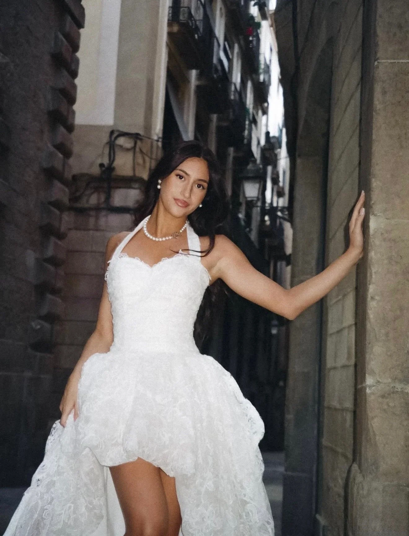 A woman in a white wedding dress standing outdoors, leaning against a wall with one arm extended, smiling softly at the camera.