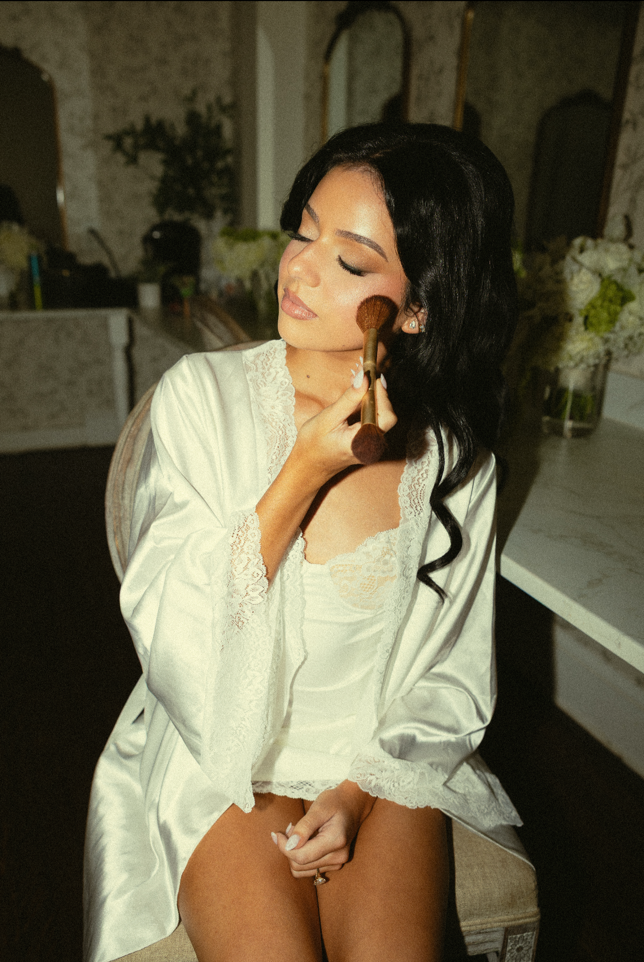 A woman with black hair, wearing a white lace-trimmed satin robe, is sitting with her eyes closed and applying makeup with a brush to her cheek.