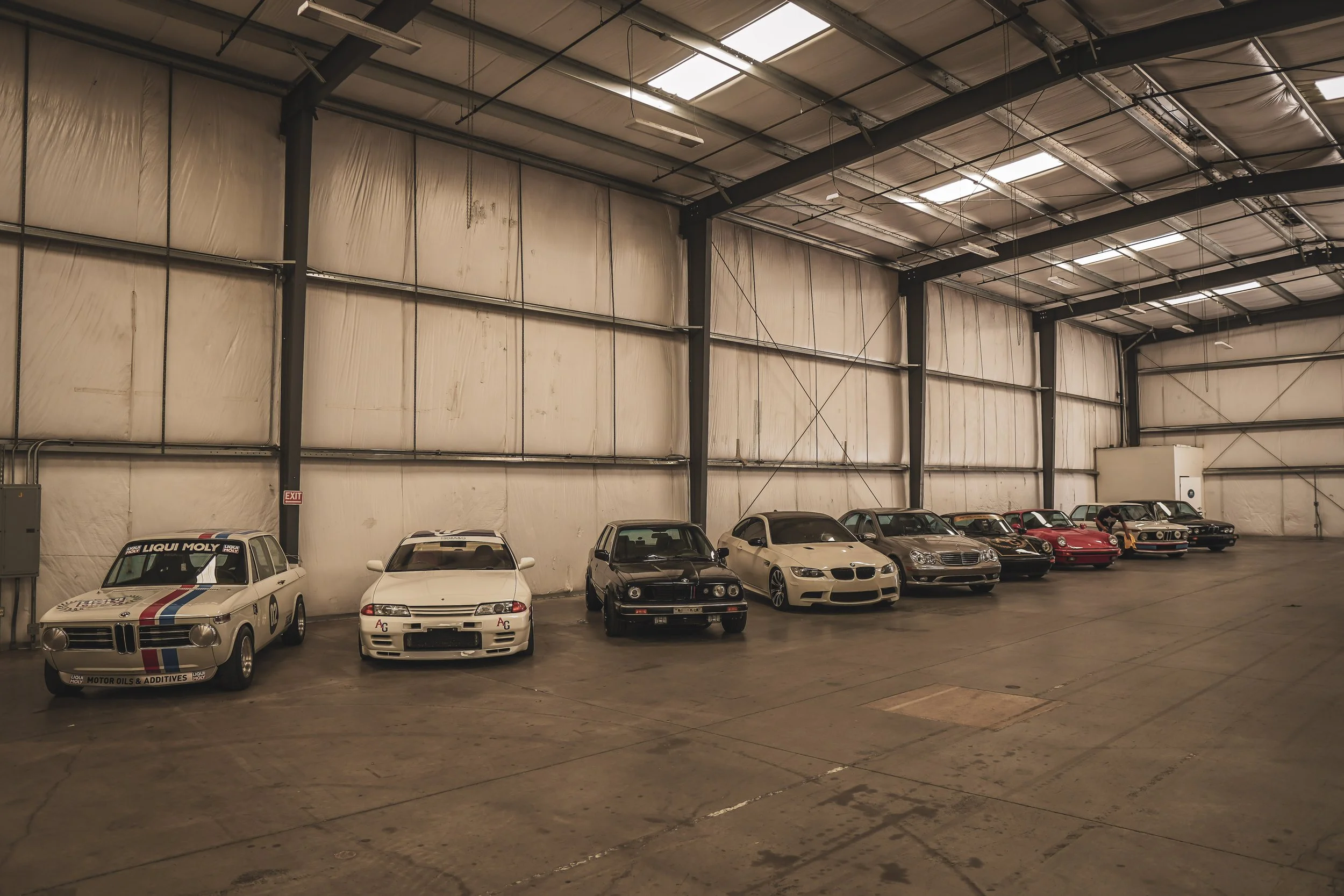 Interior of a spacious car storage warehouse with a row of classic and vintage cars parked along the wall.