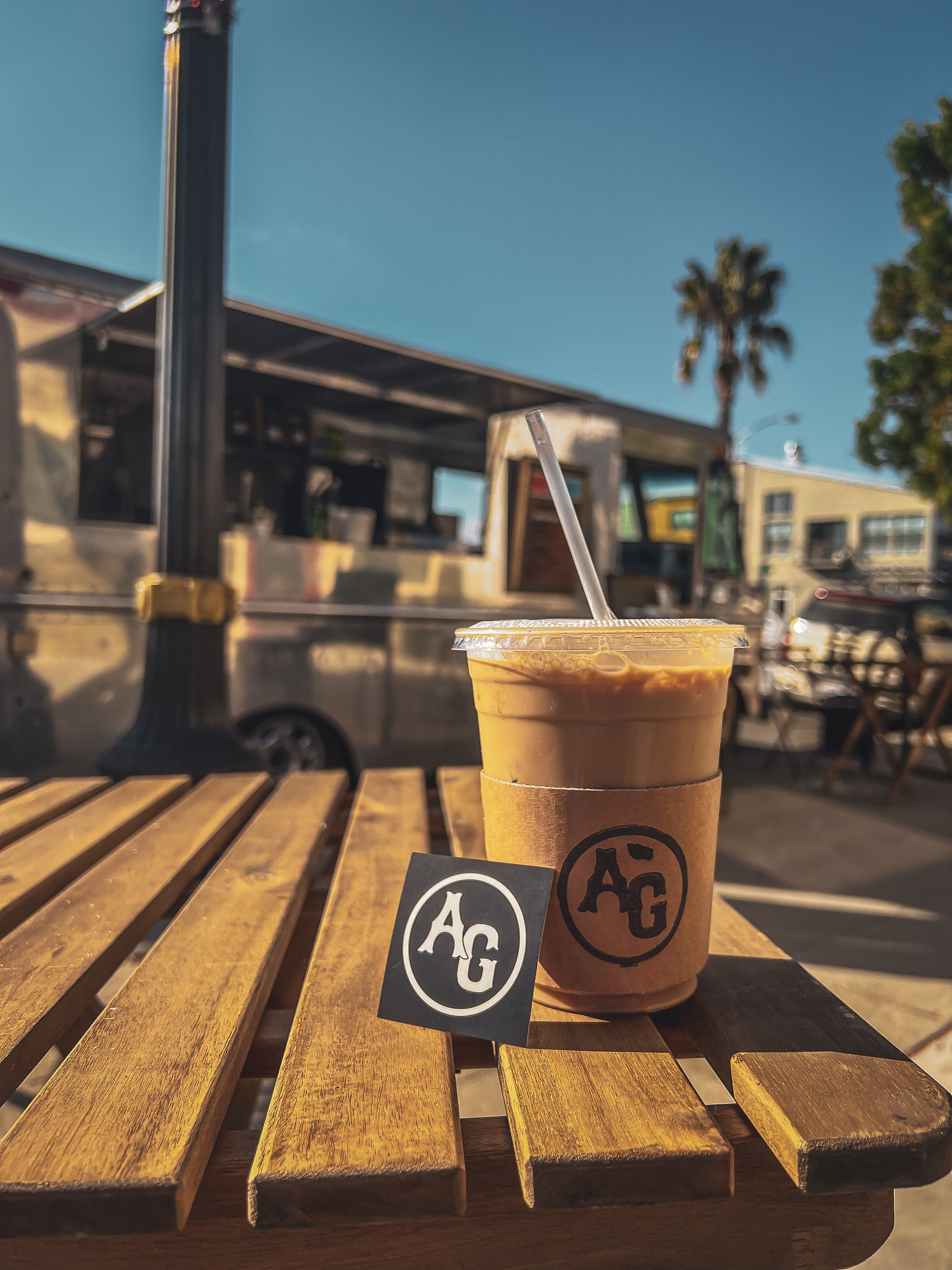 Cars and coffee with Analog and Grit on a wooden table outdoors, with a food truck and palm trees in the background.