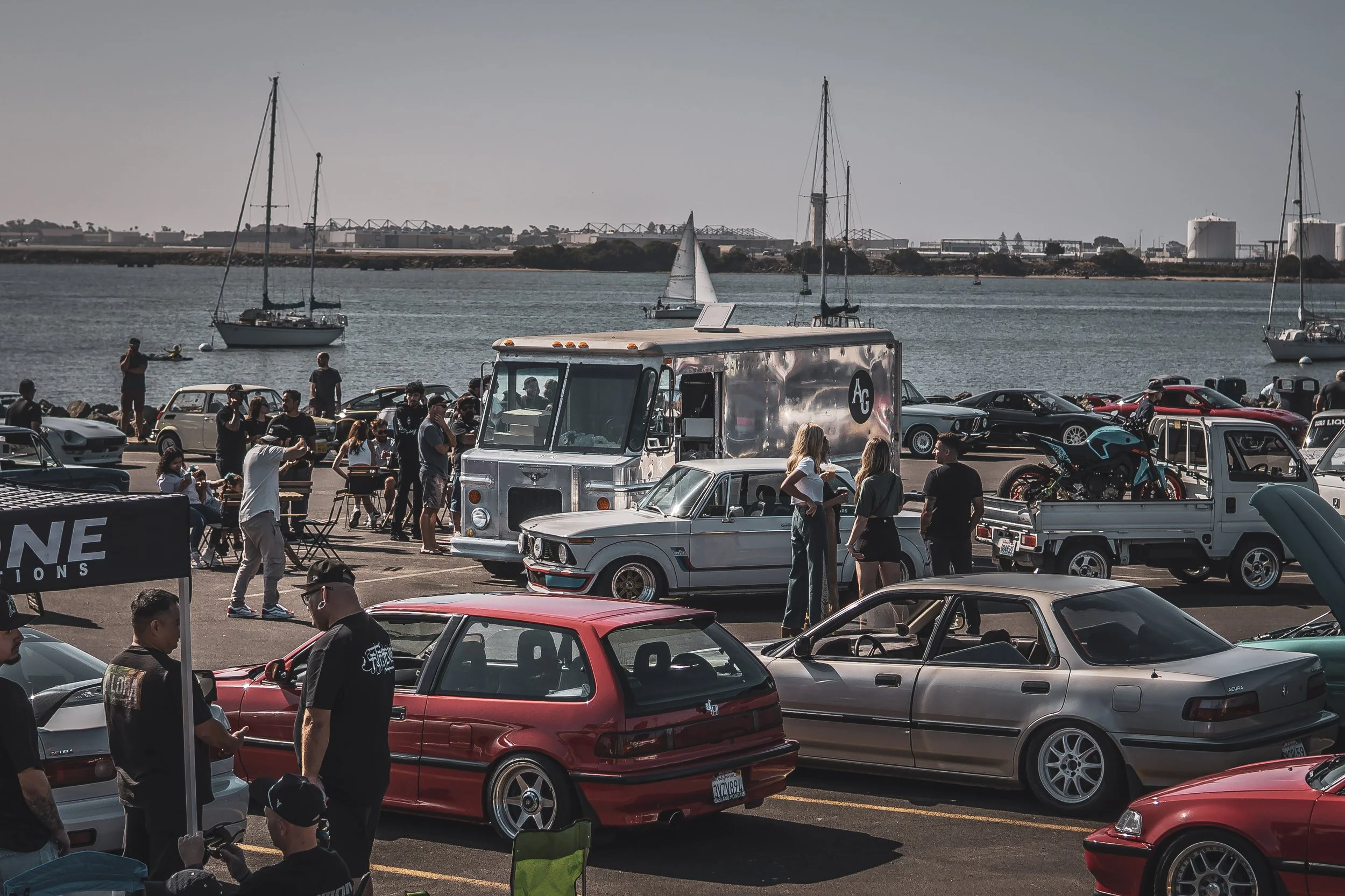 Analog and Grit car show in San Diego classic and modern cars, coffee, motorcycles and people walking and talking, sailboats in the background.