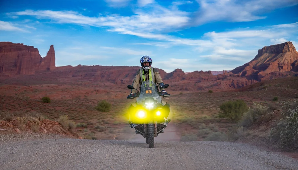A rider on a BMW with yellow lights on a gravel road among red rock buttes at sunset