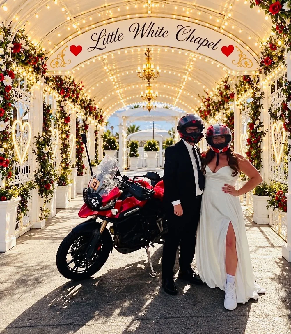 A bride and groom in motorcycle helmets beside a Triumph under the Little White Chapel sign
