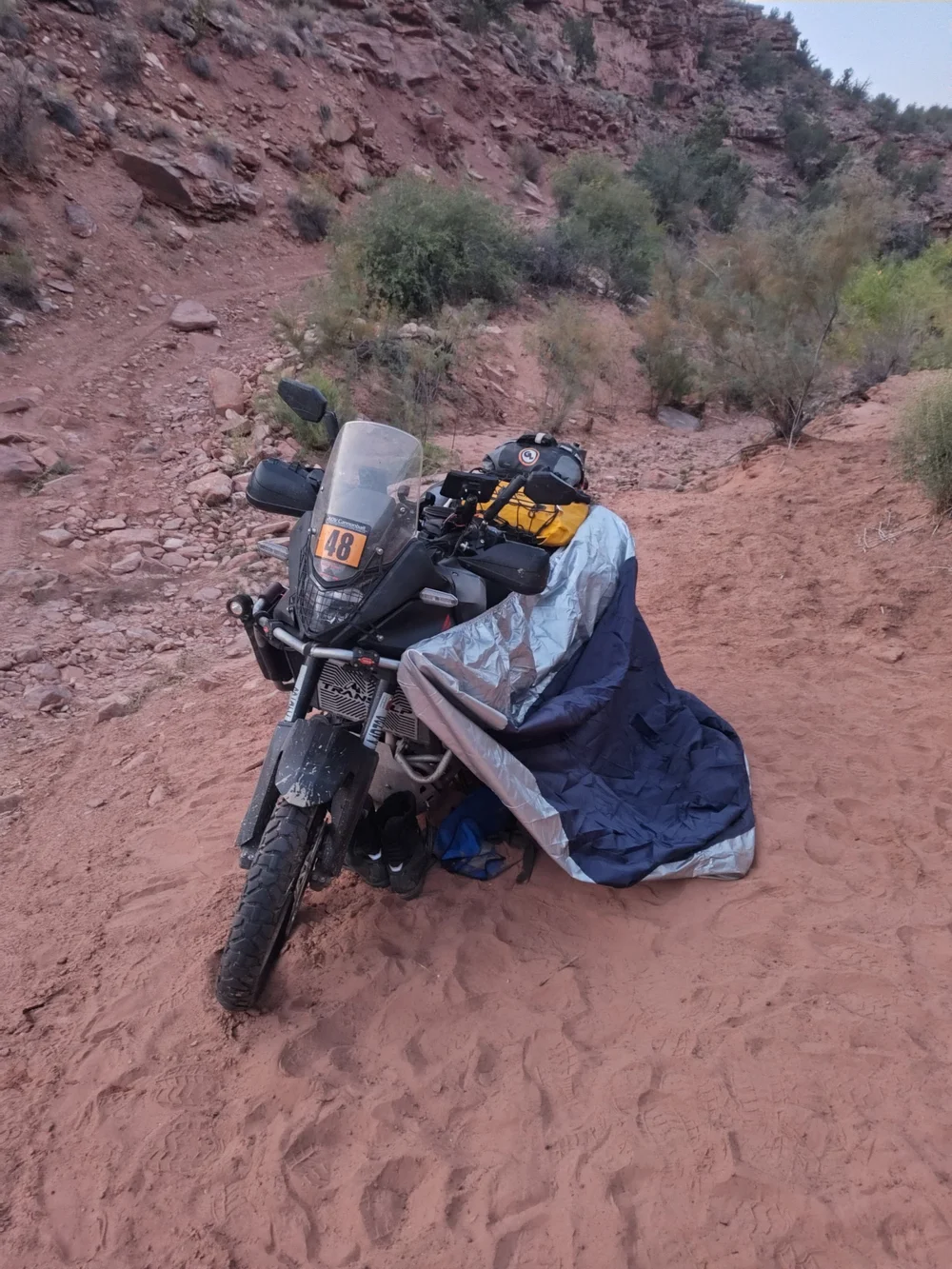 A Honda Transalp numbered 48 standing in red sand with a silver and blue motorcycle cover draped beside it as a shelter